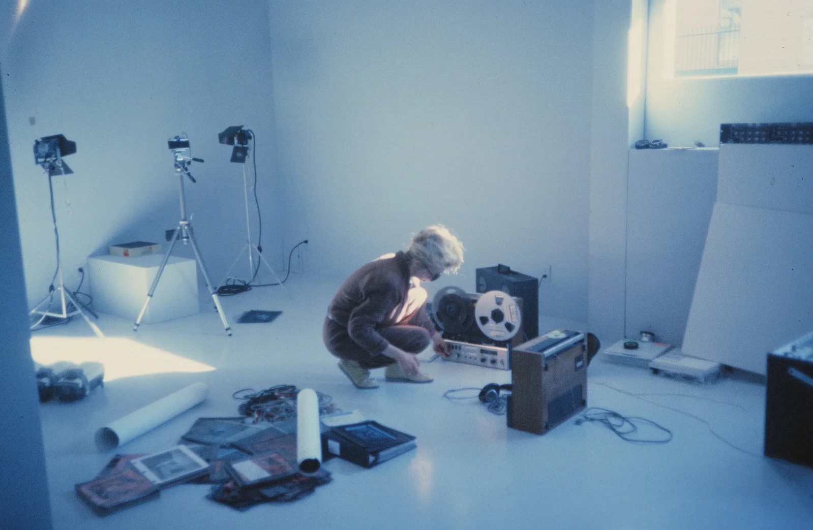 A person with short blond hair crouches in a minimalist, white-walled studio, adjusting a reel-to-reel tape machine surrounded by cables, electronic equipment, and scattered papers. Two vintage video cameras on tripods stand nearby, and soft natural light streams in from a high window, casting blue tones across the space.
