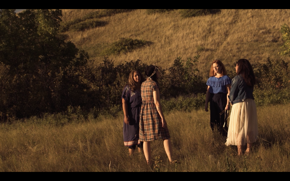Four people stand barefoot in a grassy field at golden hour, surrounded by shrubs and a hillside. They wear dresses and skirts in different styles—one in plaid with a floral crown, another in a blue embroidered dress, one in a blue off-shoulder top, and one in a white skirt with a denim jacket. The scene has a warm, cinematic atmosphere.