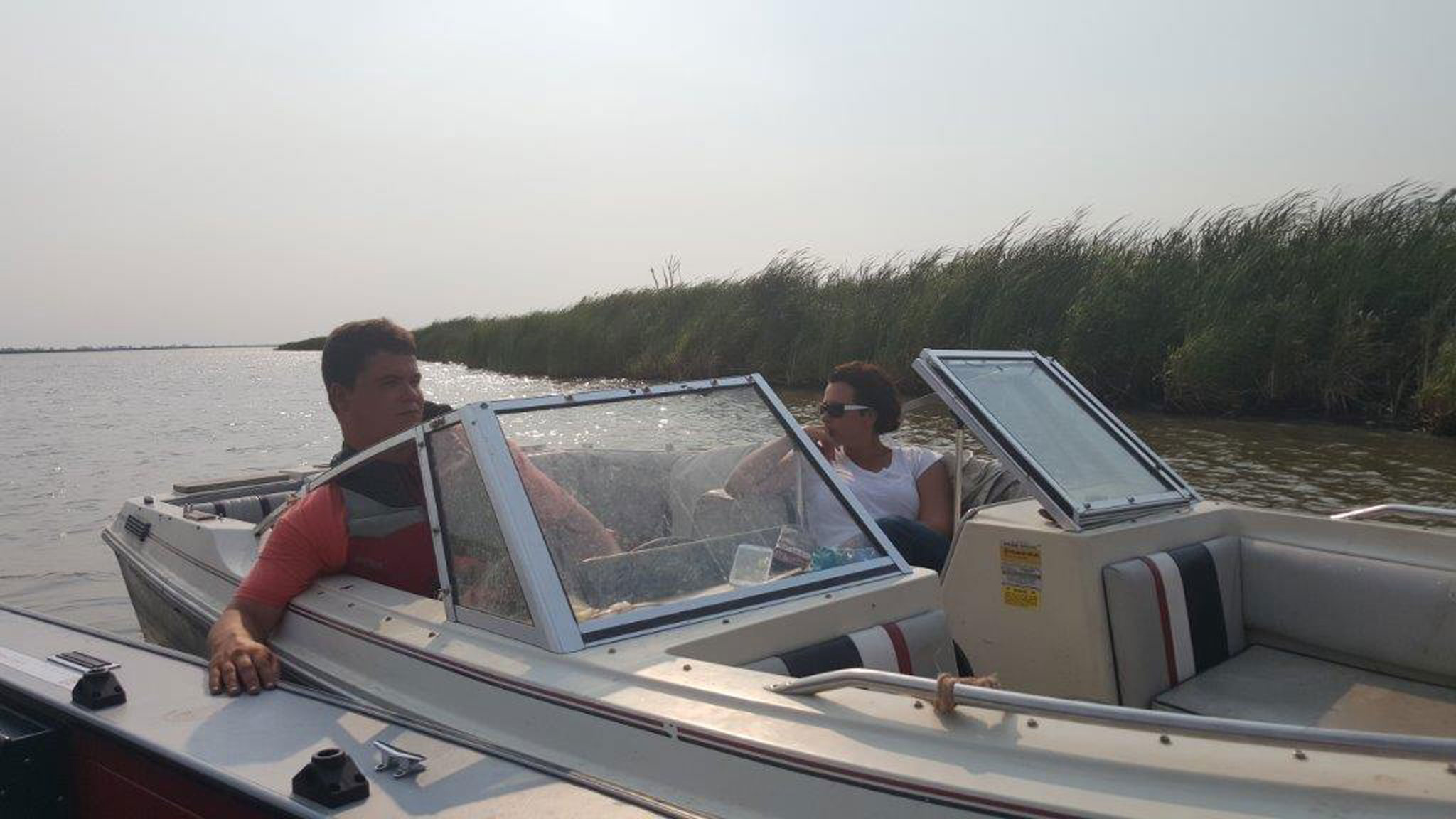 Two people sit in a small motorboat on a calm river, with tall grass lining the opposite bank. The man in front wears a red shirt and life jacket, holding the edge of another boat. The woman in the back wears sunglasses and a white shirt, resting her arm on the side and looking toward the front. The sky is hazy, casting soft light over the water.