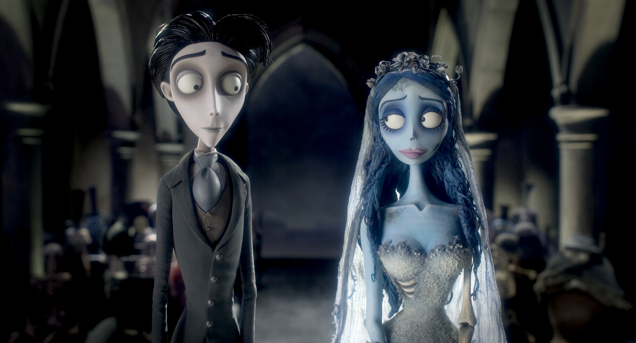 A stylized stop-motion animation of a pale groom in a grey suit and a blue-skinned bride in a tattered wedding dress and veil, standing side by side in a dimly lit gothic church. The groom looks nervously at the bride, who gazes back with a soft, melancholic expression. The background features tall stone arches and a blurred crowd of figures.