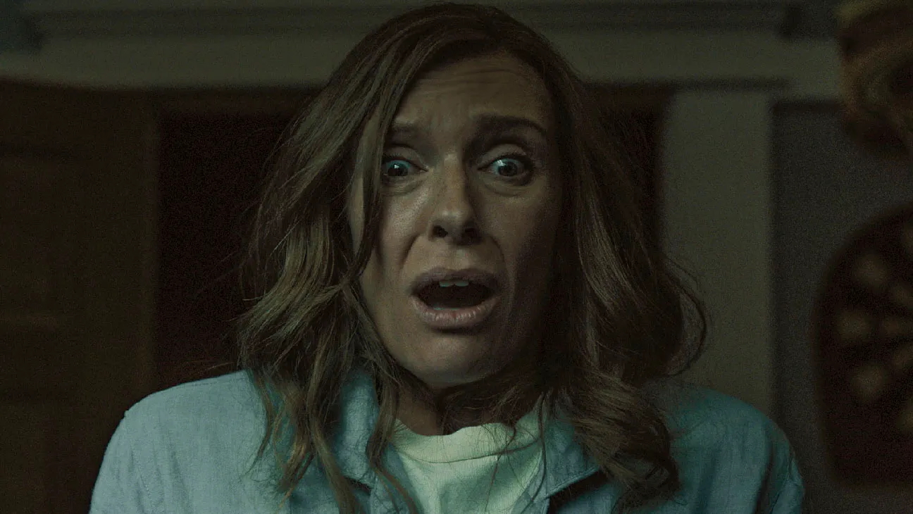 A woman with long brown hair stares forward in terror, mouth open and eyes wide in shock. She wears a pale blue robe and stands in a dimly lit interior, her expression conveying intense fear and distress.