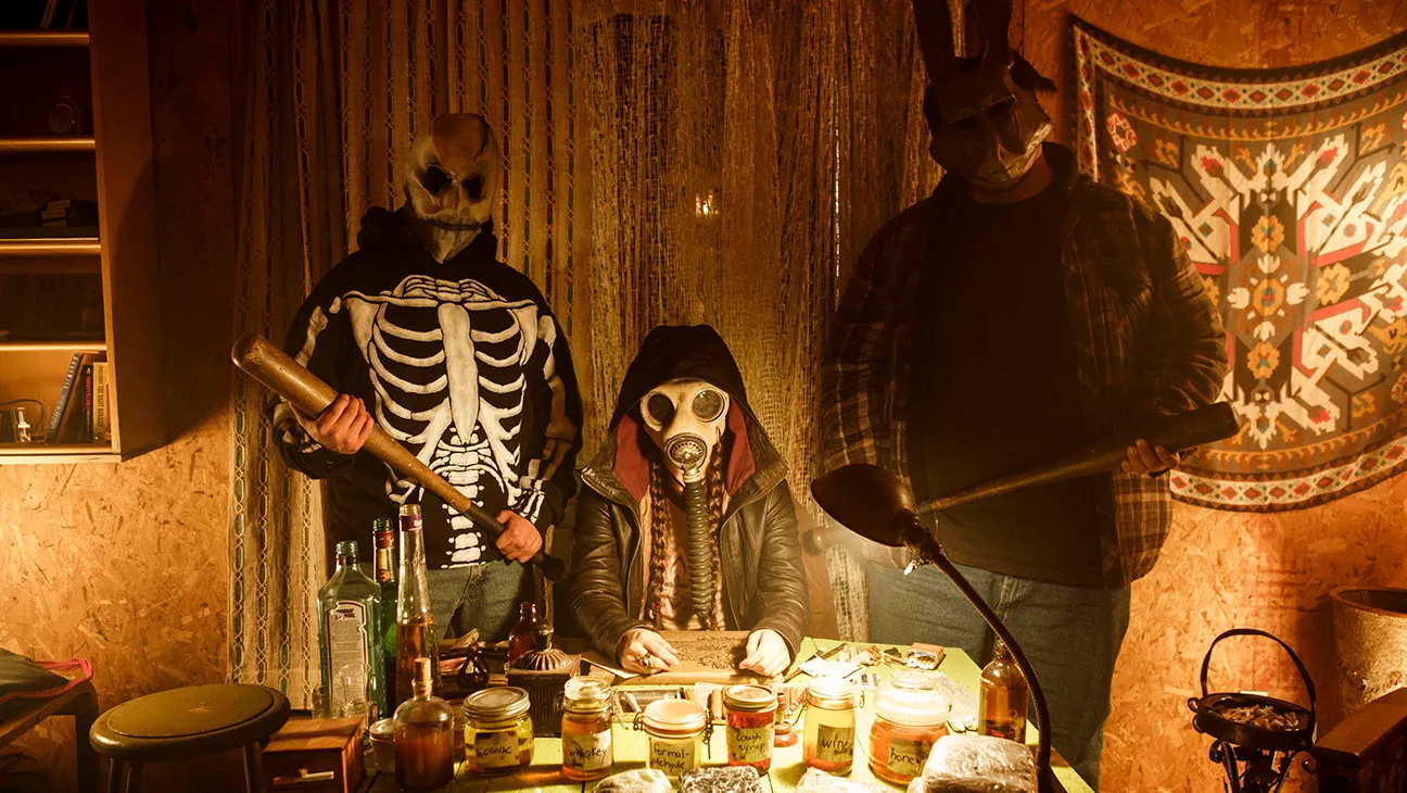 Three masked figures sit and stand around a cluttered, dimly lit table inside a wood-paneled room. The person in the centre wears a gas mask and hooded jacket, seated at the table covered in jars, bottles, and papers. On either side, two people stand holding baseball bats—one in a skeleton hoodie with a skull mask, the other in a flannel shirt with a long-eared animal mask. A patterned tapestry hangs on the wall behind them, contributing to the eerie, underground atmosphere.