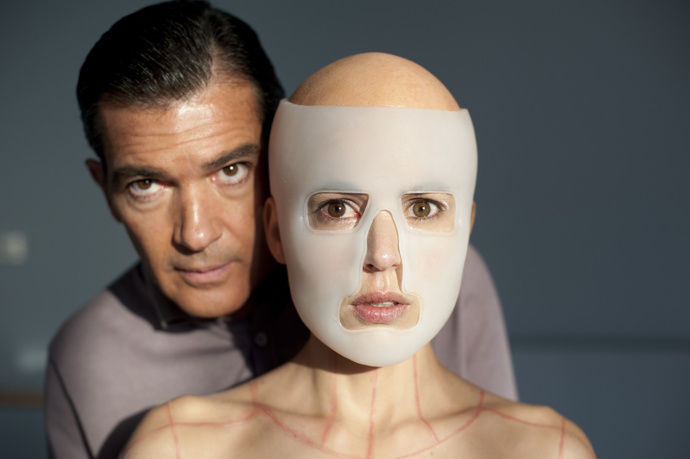 A man stands closely behind a bald woman wearing a smooth, flesh-toned medical mask that covers most of her face, revealing only her eyes, nose, and mouth. The woman’s exposed skin shows red surgical markings, and both subjects stare intensely at the camera. The lighting casts sharp shadows, enhancing the eerie and clinical atmosphere.