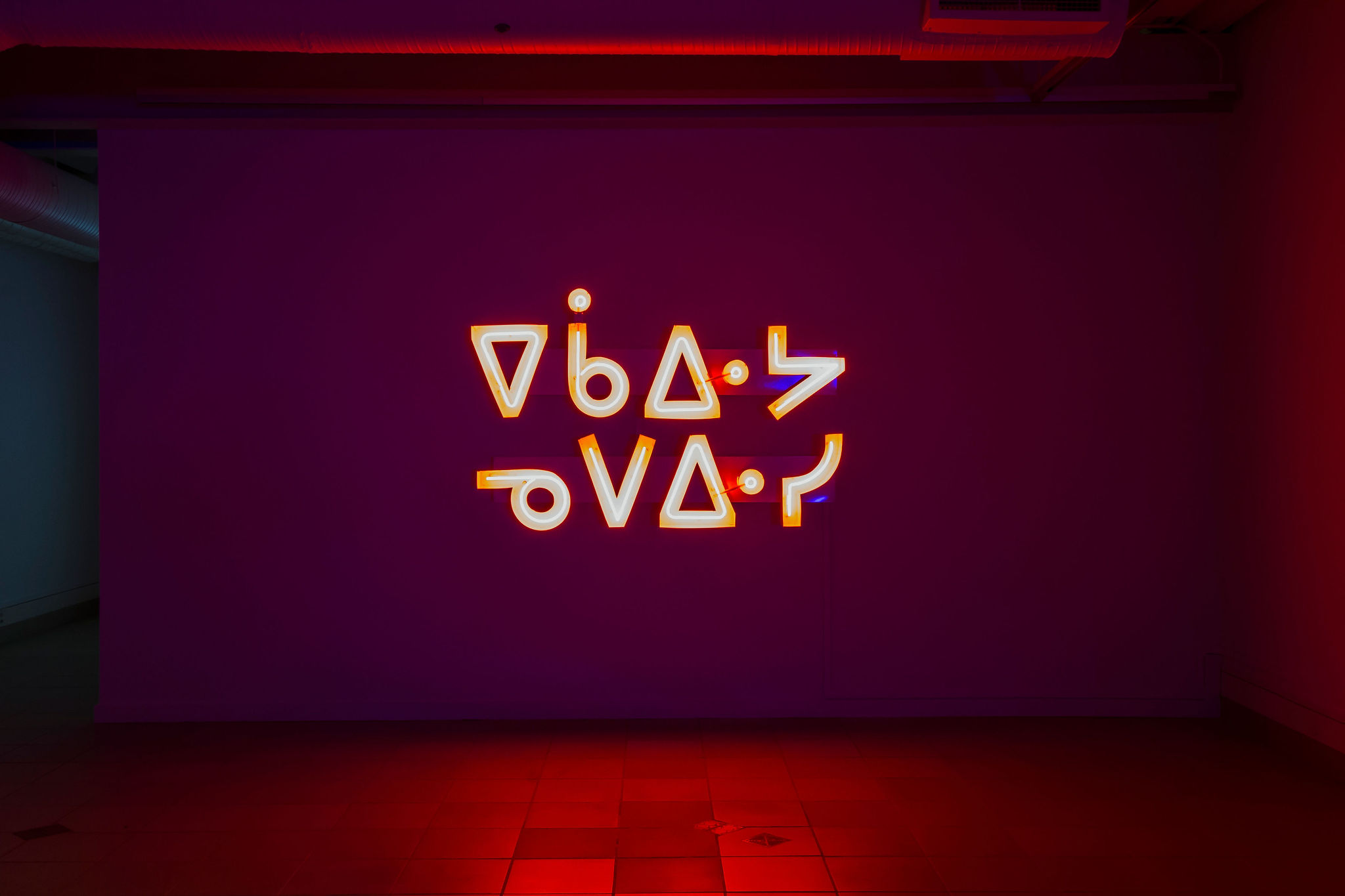 A neon sign in Indigenous syllabics glows against a deep magenta wall. The characters are outlined in bright orange and white light, casting a warm red reflection on the tiled floor. The artwork appears in a dimly lit gallery space, illuminated primarily by the vibrant signage.