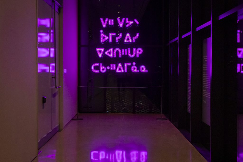 A dimly lit hallway glows with bright magenta light from a neon installation displaying stylized syllabic characters. The glowing text reflects on the polished floor, while the surrounding walls and glass surfaces catch the vivid purple hues.