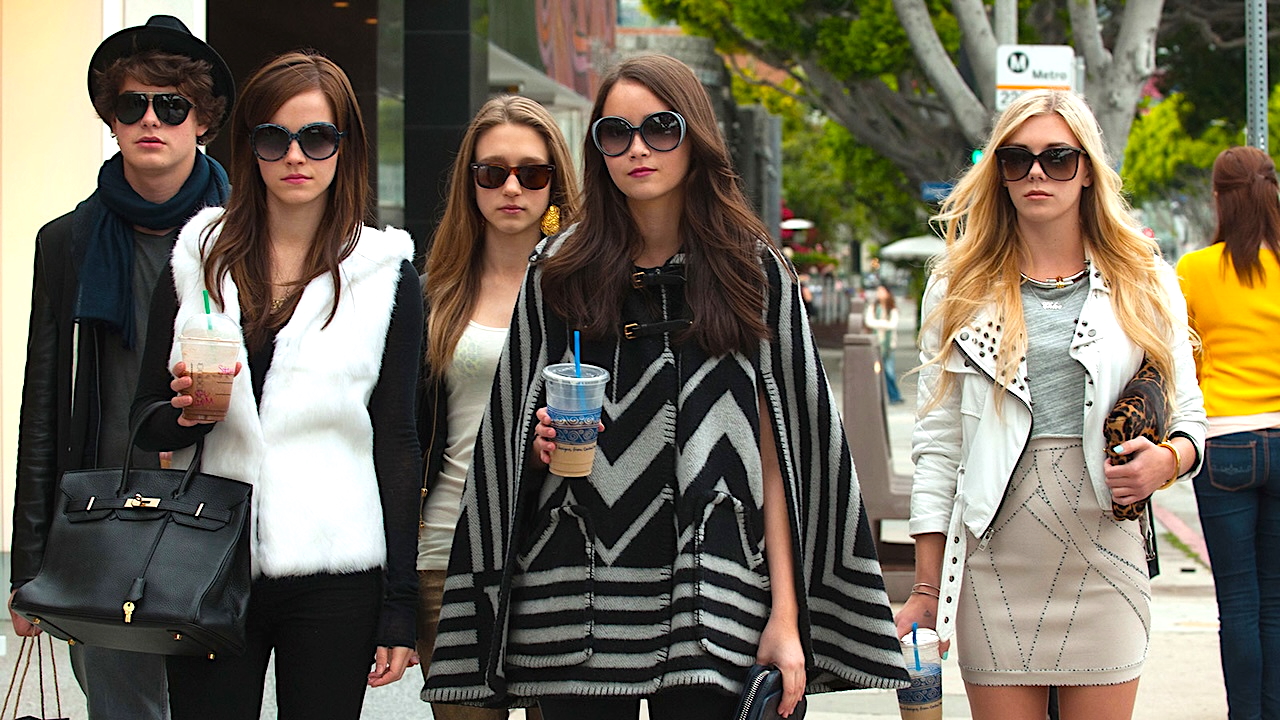 BLING RING_Remai A stylish group of five young adults walk confidently down a city sidewalk, all wearing oversized sunglasses and fashionable outfits. Two carry iced drinks, and one holds a large black handbag. Trees, pedestrians, and storefronts line the background, evoking an upscale urban setting.