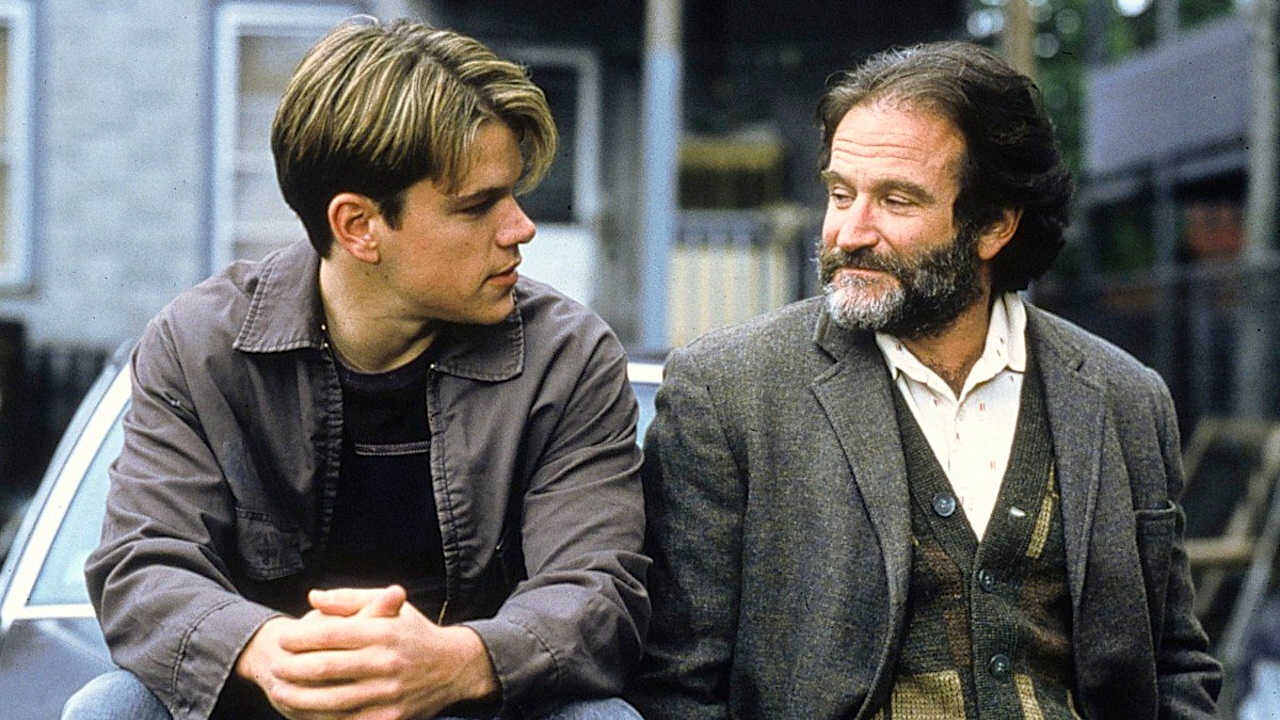 Matt Damon and Robin Williams sit on a bench in a heartfelt moment from the film Good Will Hunting. Damon, playing Will, wears a grey jacket and looks intently at Williams, who plays therapist Sean Maguire. Williams, in a worn blazer and cardigan, returns the gaze with a gentle, knowing expression. They sit outdoors, backed by blurred buildings and muted urban scenery, underscoring the film’s emotional and introspective tone.