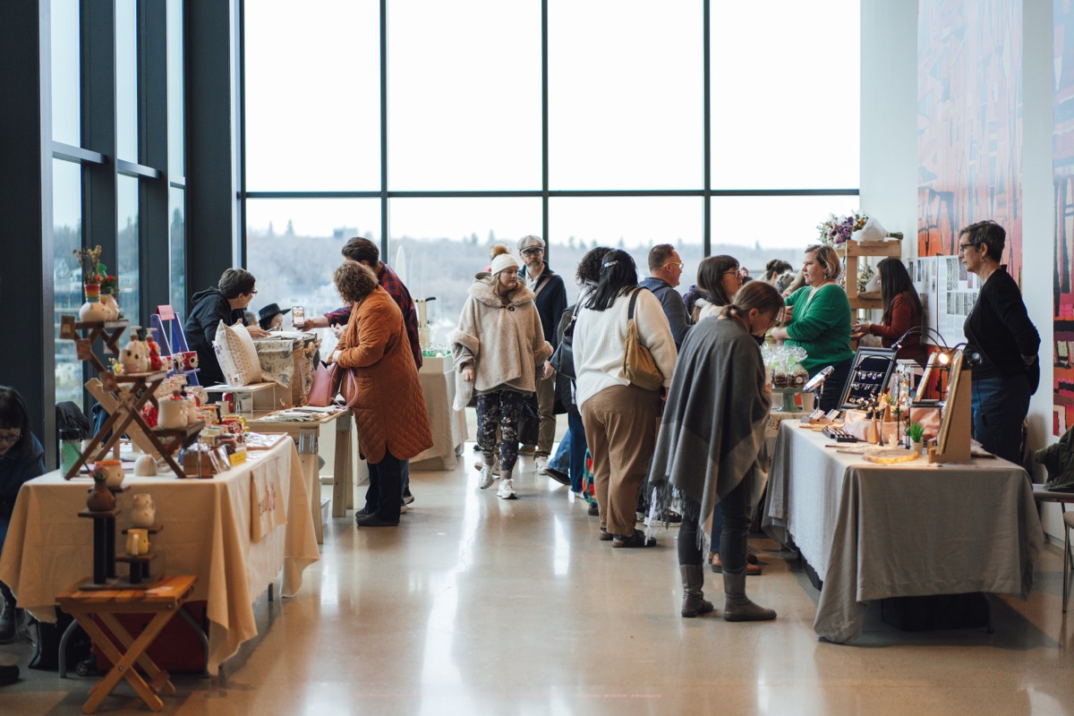 Tiger Lily Market_Remai Modern A bustling indoor market with vendors and visitors interacting at tables displaying handmade goods, art, and crafts. The space has large floor-to-ceiling windows letting in natural light and offering a view of trees and the river outside.