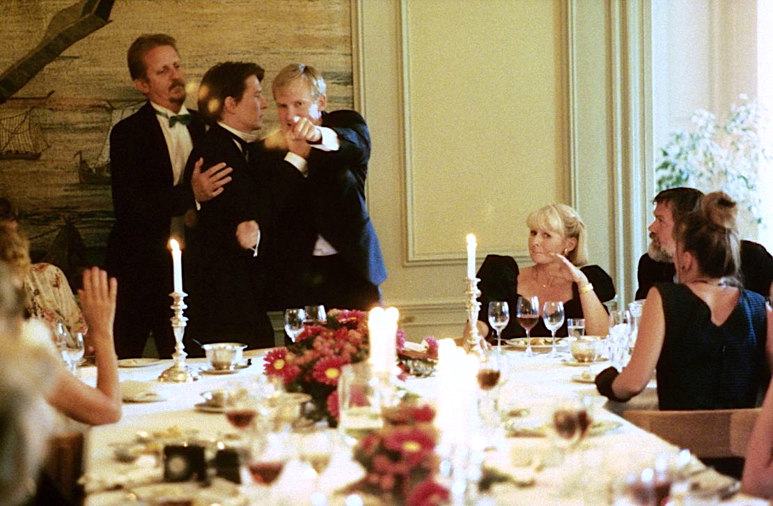 A tense moment unfolds at an elegant dinner party. Three men in tuxedos stand at one end of a long, candlelit table—one man aggressively pointing, another trying to restrain him. Guests seated around the table, dressed formally, watch the confrontation with concern and discomfort. The room is softly lit with ornate decor, floral arrangements, and half-filled wine glasses.