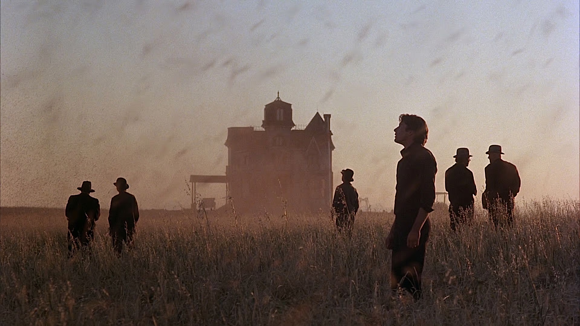 Silhouetted figures in dark clothing walk through a field of tall grass at dusk, a dramatic Victorian-style house looming in the hazy background. The sky is streaked with the movement of a locust swarm, giving the scene a sense of eerie stillness and foreboding.