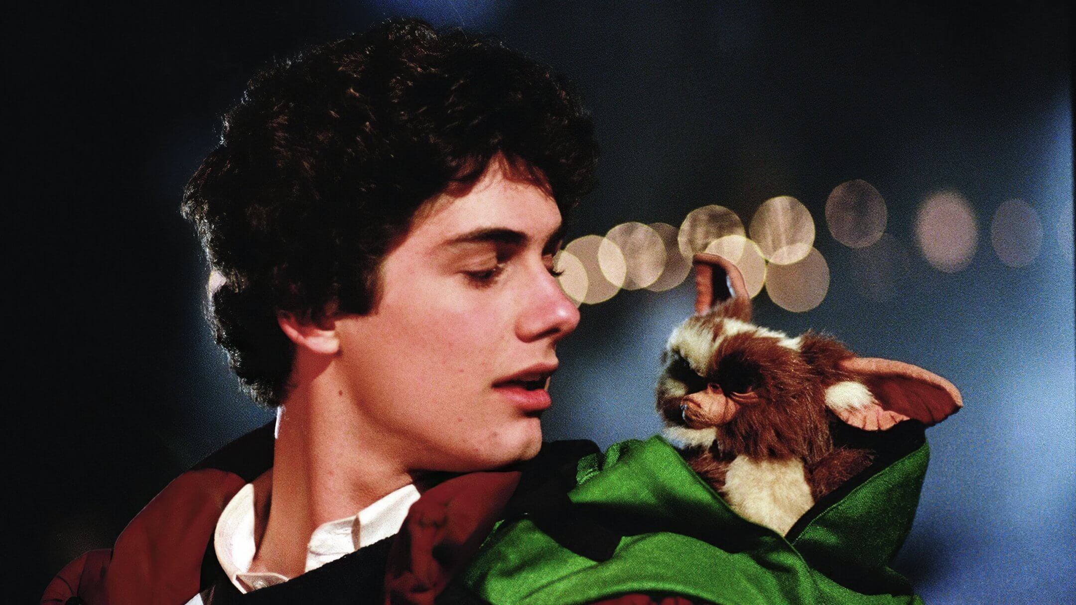 A young man with curly dark hair looks down affectionately at a small furry creature peeking out of his green jacket. The creature, with large ears and wide eyes, resembles a mix between a gremlin and a teddy bear. Soft bokeh lights glow in the background, adding a warm, magical atmosphere to the scene.