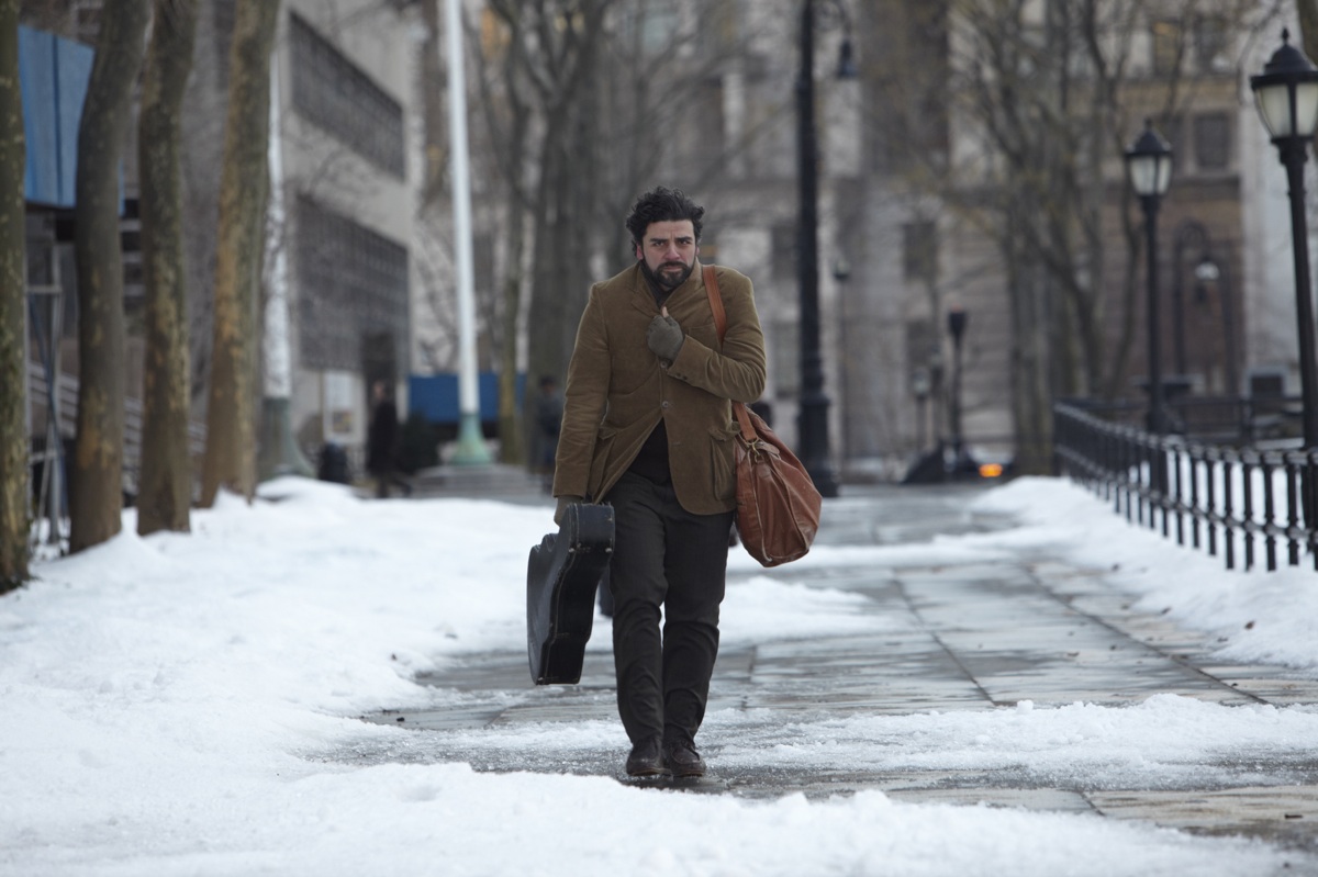 A man with disheveled hair and a beard walks alone down a snow-covered sidewalk in winter, clutching a guitar case in one hand and a worn leather bag over his shoulder. He wears a brown corduroy jacket and dark gloves, his expression tired and introspective. Bare trees and classic streetlamps line the path, with tall buildings in the background.