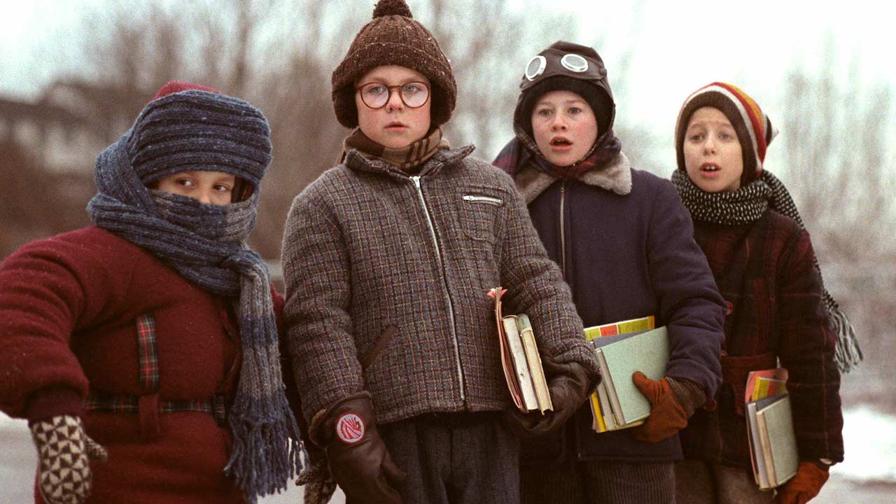Four bundled-up children stand outdoors in winter, holding books and notebooks against a snowy backdrop. The child on the left is wrapped tightly in a scarf with only their eyes visible, while the others wear thick coats, hats, and gloves. One child wears round glasses and a serious expression; another sports aviator-style headgear. They appear surprised or concerned, evoking a nostalgic, vintage winter scene.
