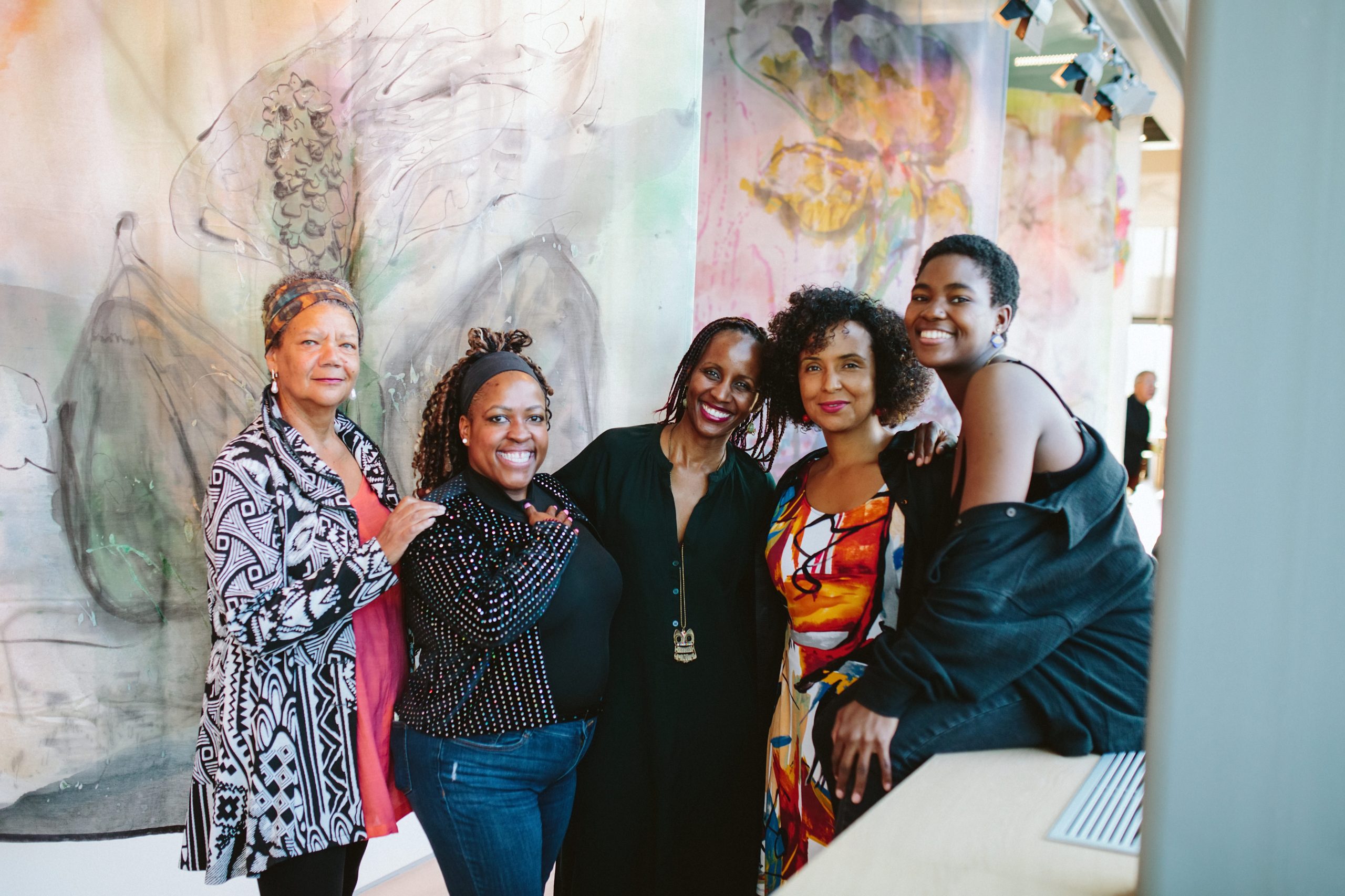 Five women stand closely together indoors, smiling warmly at the camera. They pose in front of large, softly colored abstract textile artworks with floral and organic forms. The women wear a mix of patterned and solid-color clothing, and one leans casually against a ledge at the edge of the frame.
