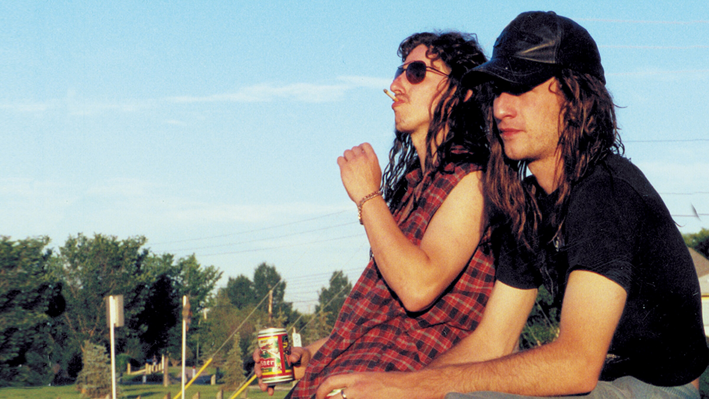 Two men with long hair sit outdoors under a clear blue sky, leaning against a low surface. One wears sunglasses and a sleeveless plaid shirt, holding a can of beer and a cigarette, while the other wears a black cap and dark T-shirt. Trees, power lines, and roadside elements appear in the background.