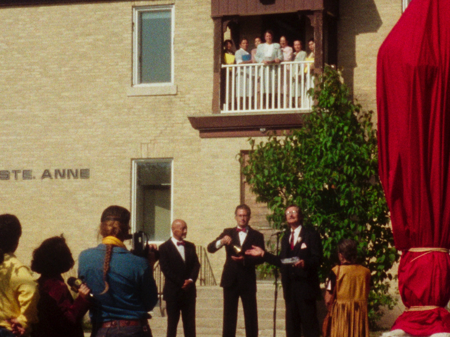 In a sunlit courtyard outside a brick institutional building, three men in dark suits stand at microphones addressing a small crowd. One gestures with an object in his hands while another appears to be speaking. Onlookers gather at ground level, and several people—mostly women and children—watch from a balcony above. At the right edge of the frame, an object draped entirely in red fabric partially obscures the scene.