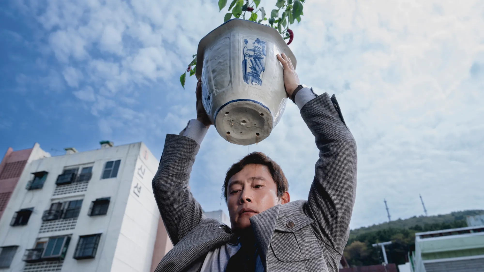 A man stands outdoors beneath a bright, cloudy sky, lifting a large ceramic plant pot above his head with both hands. A small leafy plant grows from the pot. Apartment buildings and a hillside appear in the background, and the low-angle view emphasizes the weight and tension of the moment.