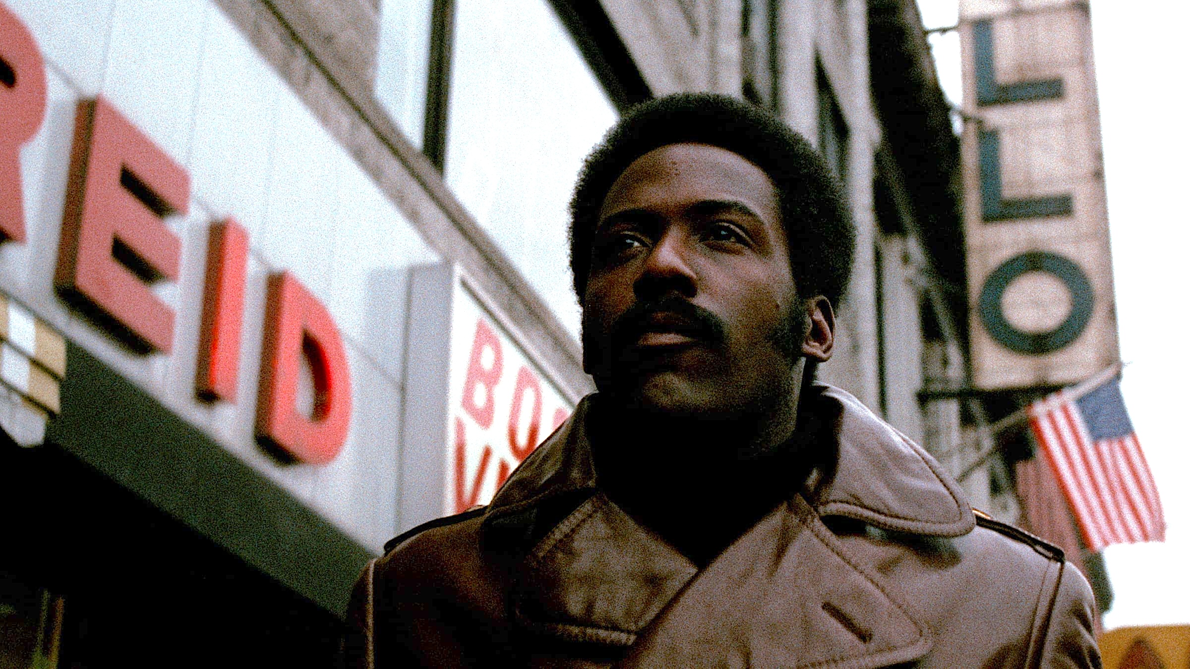 A man walks along a city street, seen from a low angle, wearing a tan coat over a dark turtleneck. He looks ahead with a calm, focused expression. Behind him, tall buildings line the street, with large storefront signage and a vertically hanging American flag visible.