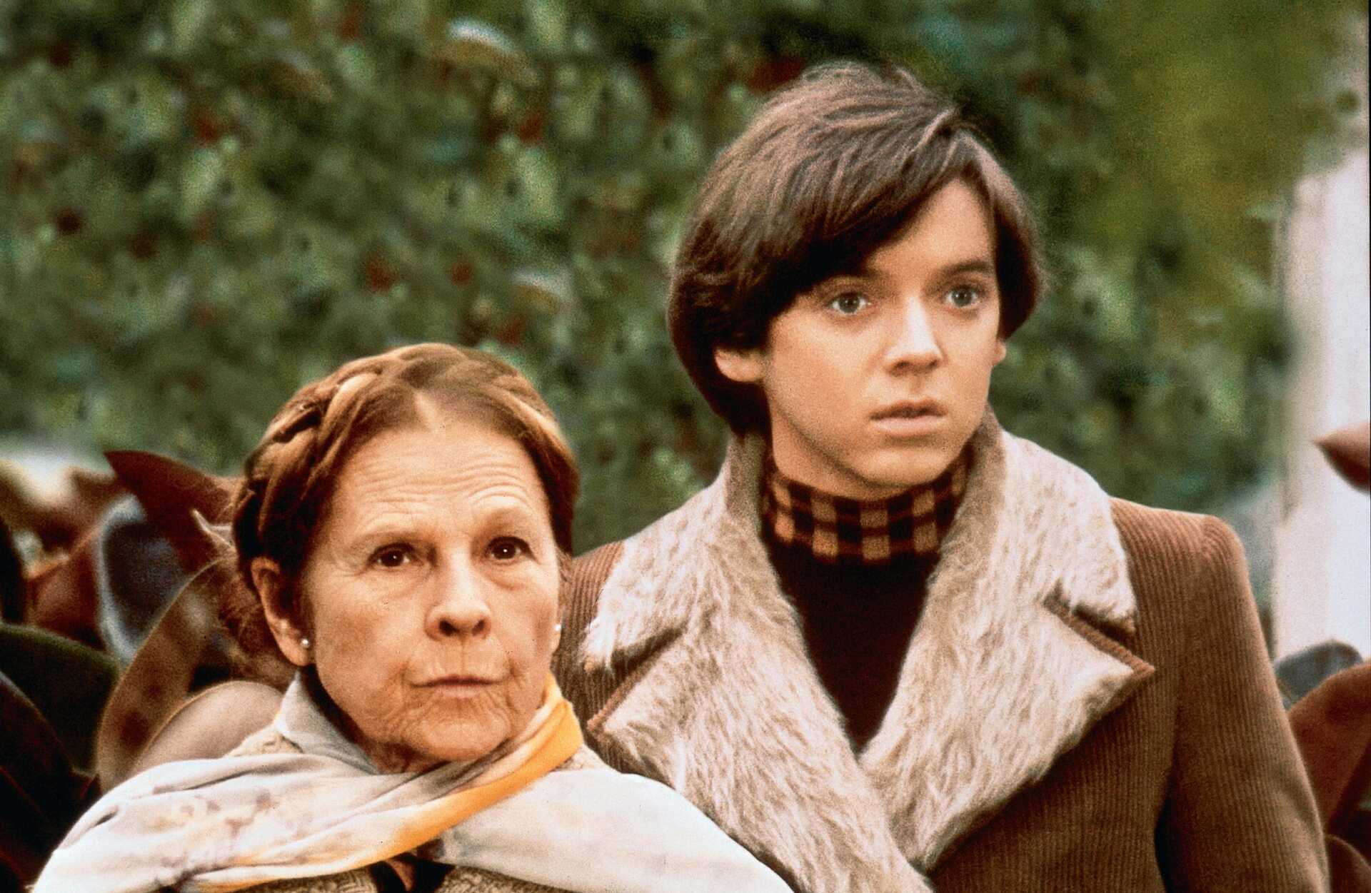 An older woman and a young person stand close together outdoors, both looking off to the side with concerned expressions. The older woman wears a light scarf and coat; the younger person wears a brown jacket with a shearling collar and a checkered turtleneck. Green foliage fills the background.