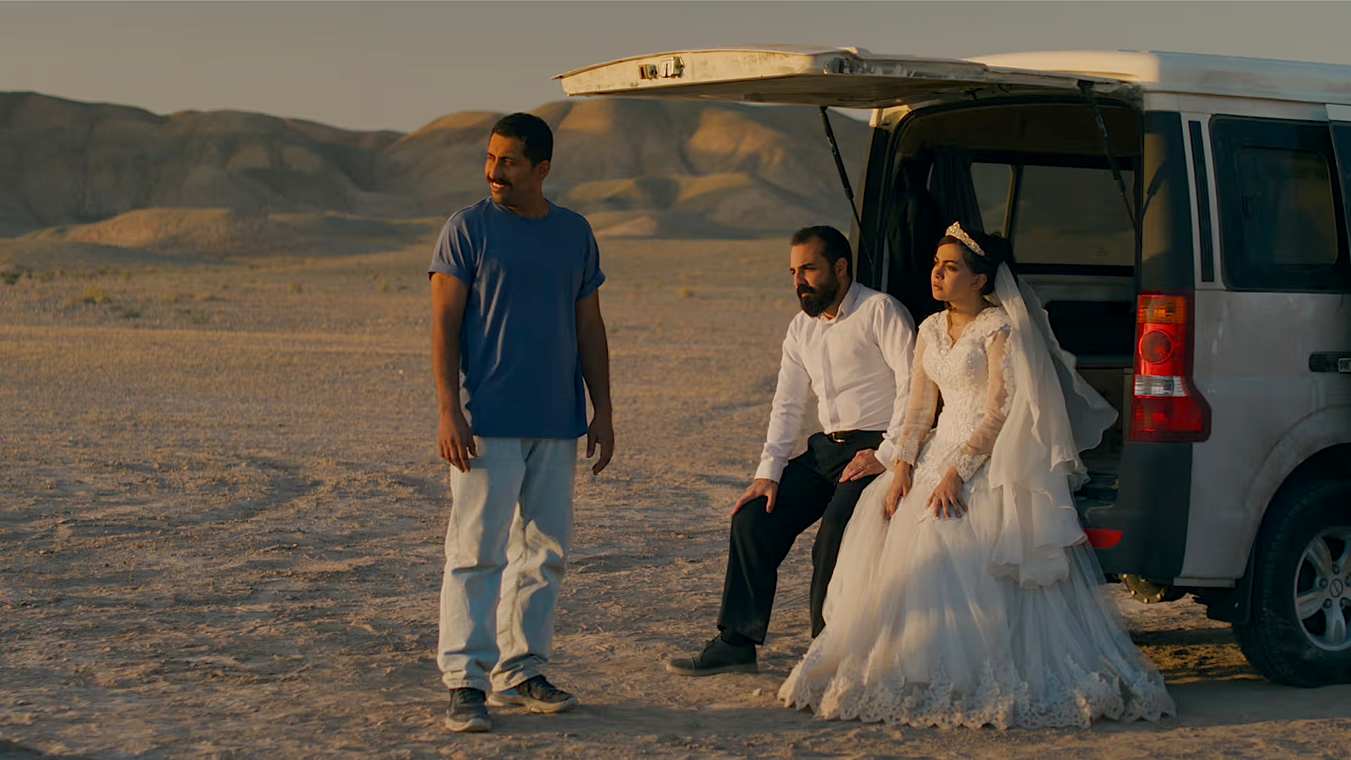In a wide desert landscape at golden hour, a bride and groom sit side by side on the open tailgate of a silver van. The bride wears a white lace wedding dress and veil, while the groom is dressed in a white shirt and dark trousers. A third man in a blue T-shirt and light pants stands nearby, looking off into the distance. Low, rocky hills stretch across the background.