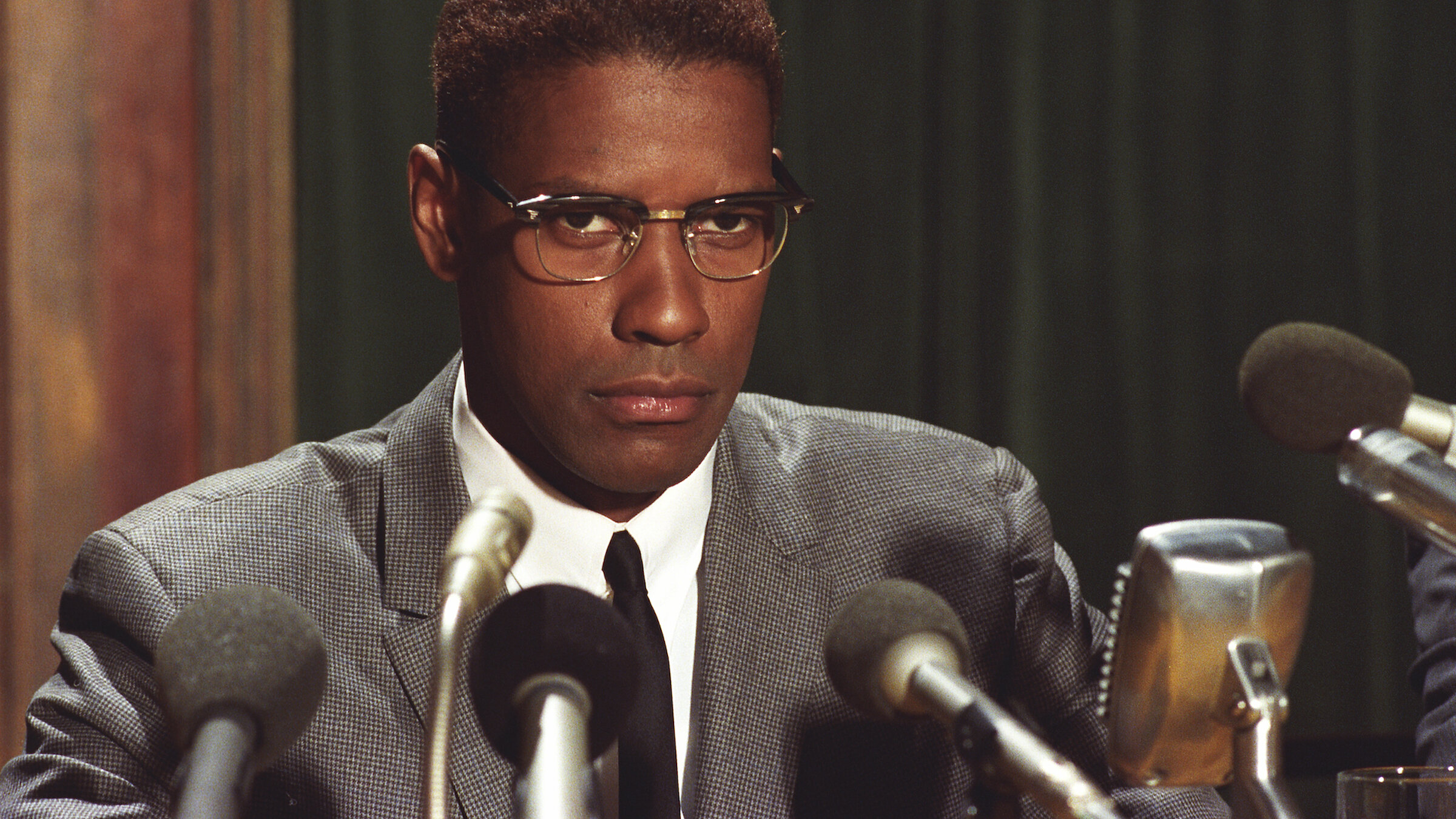 A man in a gray suit and black tie sits at a table facing several microphones, wearing rectangular eyeglasses and looking forward with a serious, focused expression. The microphones crowd the foreground, suggesting a press conference or public address.