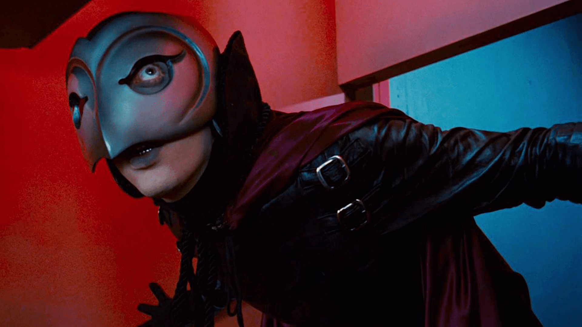 A masked figure wearing a stylized bird-like helmet and dark leather clothing leans forward dramatically in a corridor. Red and blue lighting washes over the scene, creating a bold, theatrical contrast.