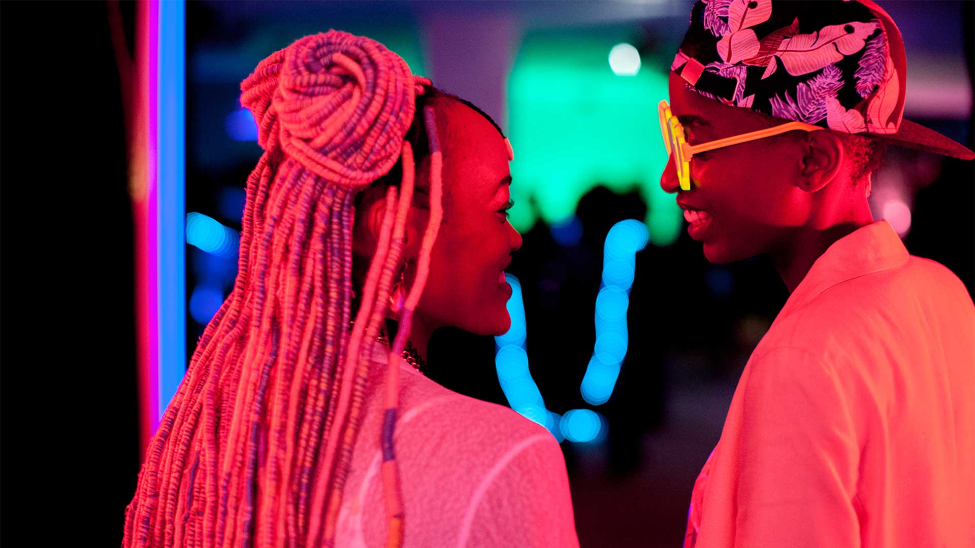 Two people face each other closely in a dark, lively indoor setting illuminated by vivid pink, blue, and green lights. One has long, light-colored braids styled into a bun, while the other wears bright yellow glasses and a patterned cap. Both appear to be smiling and engaged in conversation, with blurred lights and figures in the background.