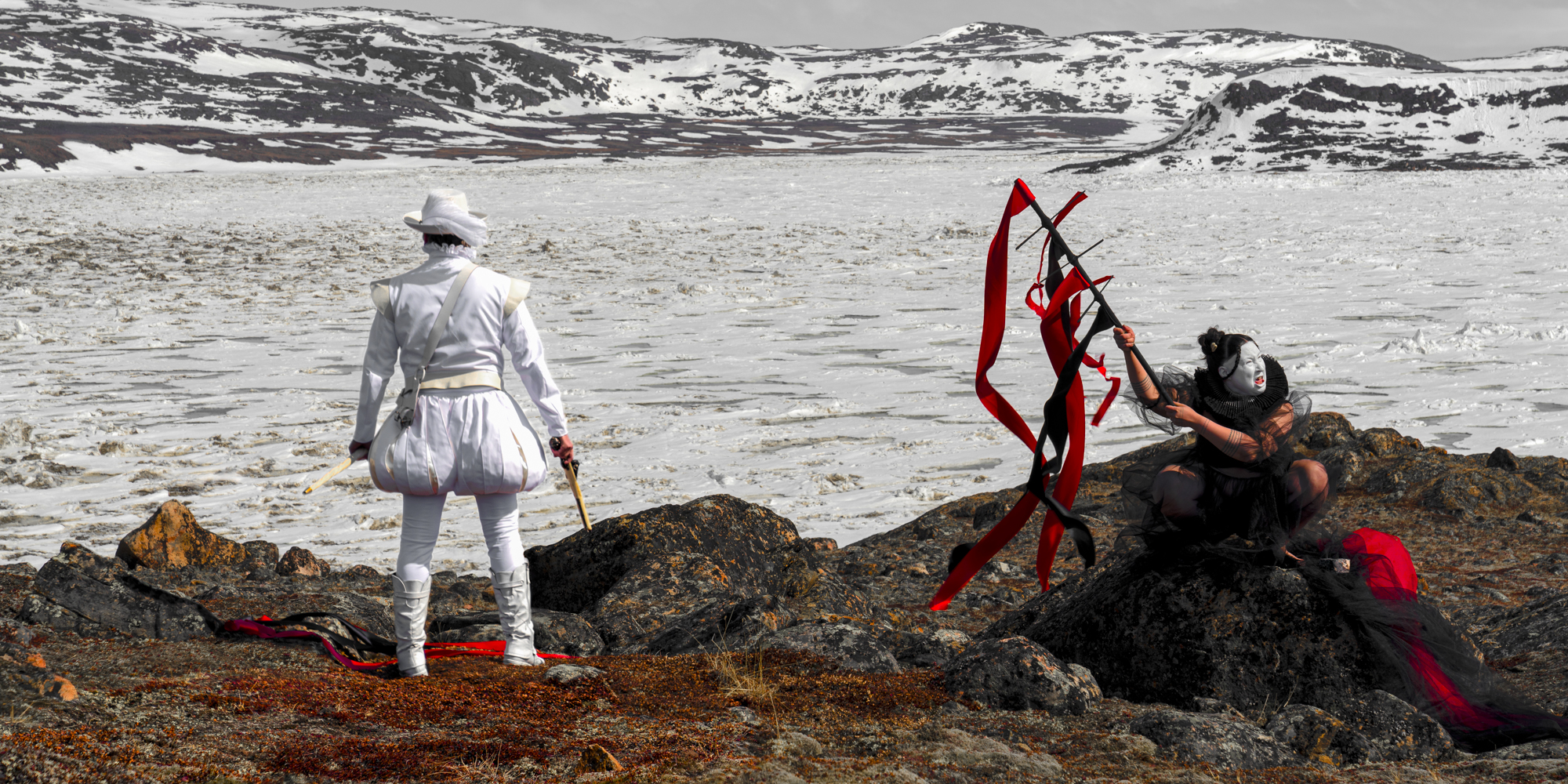 Two figures stand on dark rocks at the edge of a frozen or icy body of water, with snow-covered hills in the background. One figure, dressed entirely in white, faces the landscape, while the other, dressed in black and red with a face painted white, raises a stick adorned with long red ribbons.