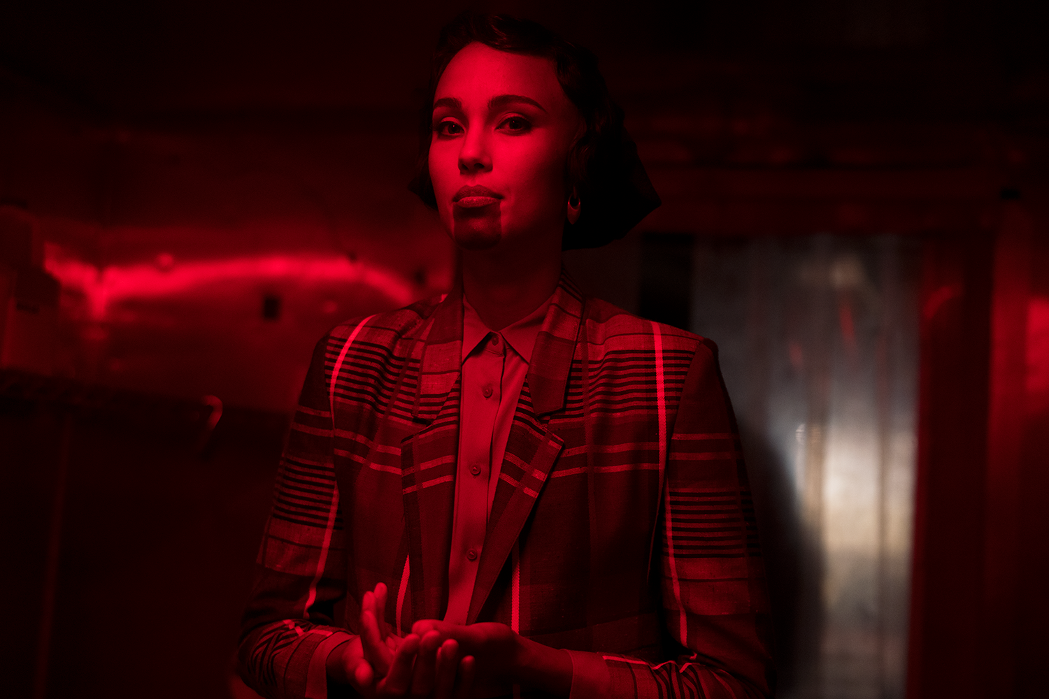 A person stands indoors under dramatic red lighting, wearing a patterned blazer over a buttoned shirt, hands gently clasped at chest height, looking forward with a calm, focused expression; the background is dim and softly lit, suggesting a gallery or performance space.