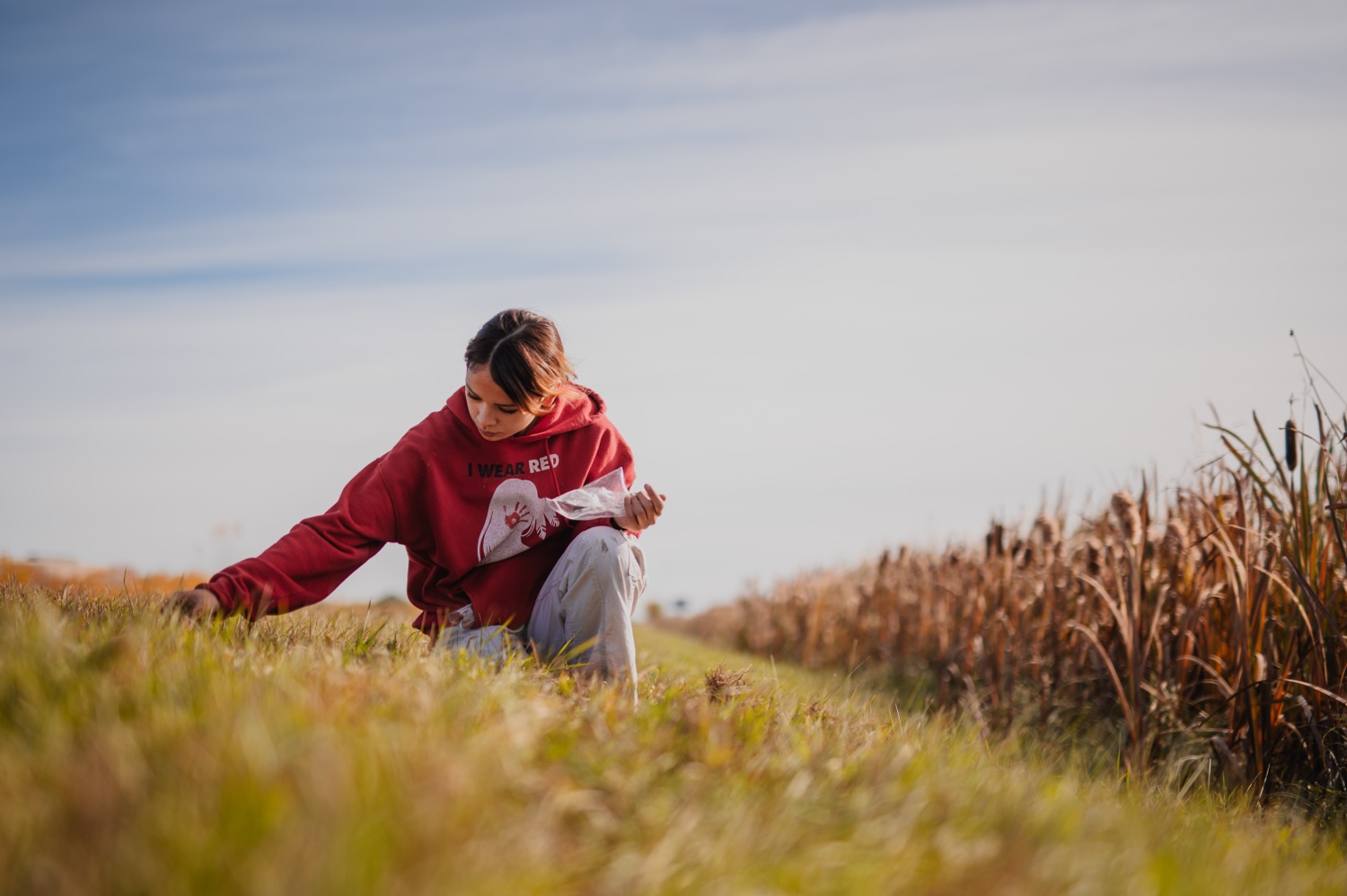 A person in a red hoodie kneels in a grassy field, planting or examining small plants while holding a plastic bag, with tall dried vegetation and open sky in the background.