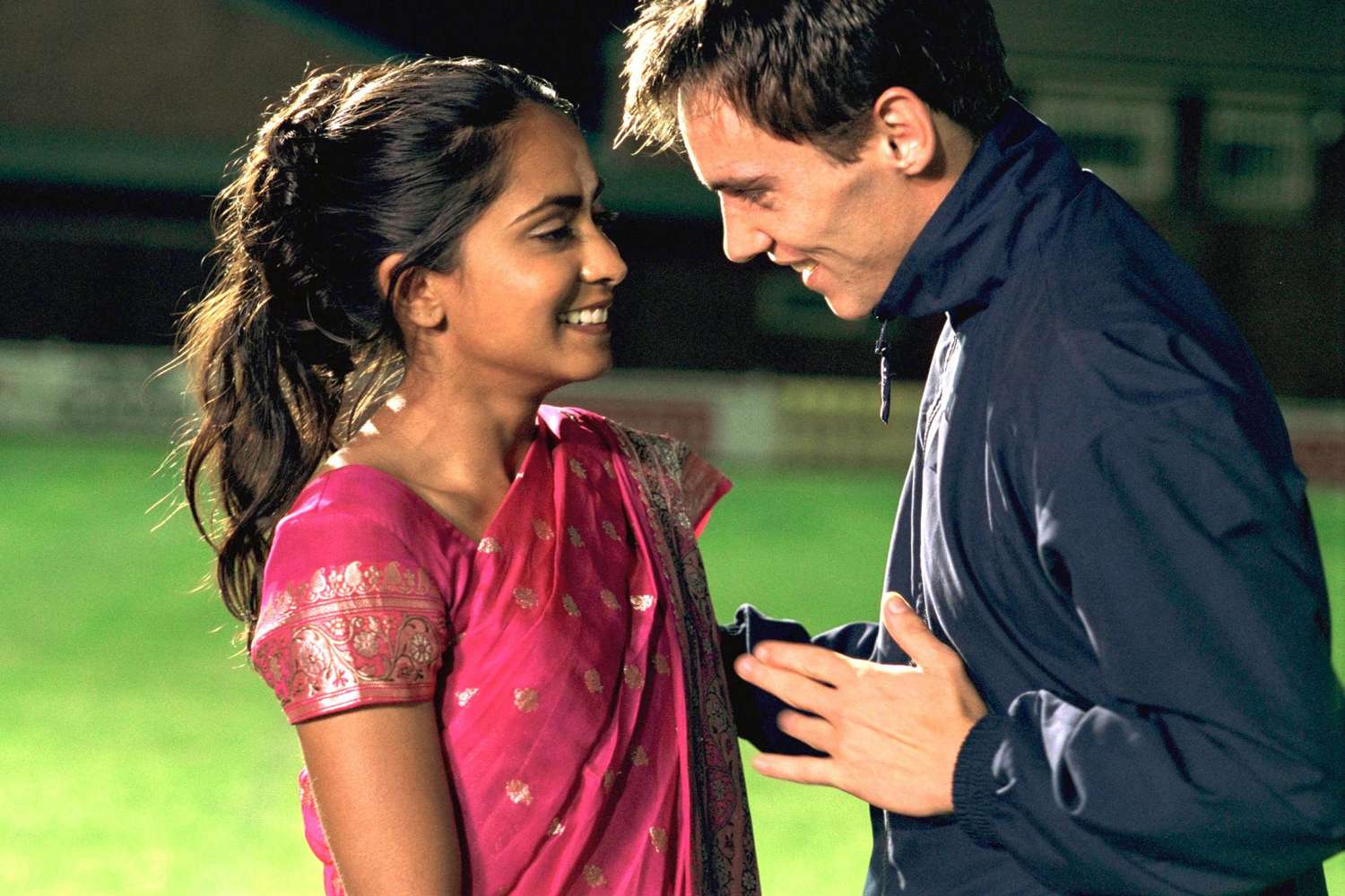 Bend It Like Beckham A young woman in a pink embroidered sari and a man in a dark zip-up jacket face each other closely on what appears to be a sports field at night, both smiling warmly.