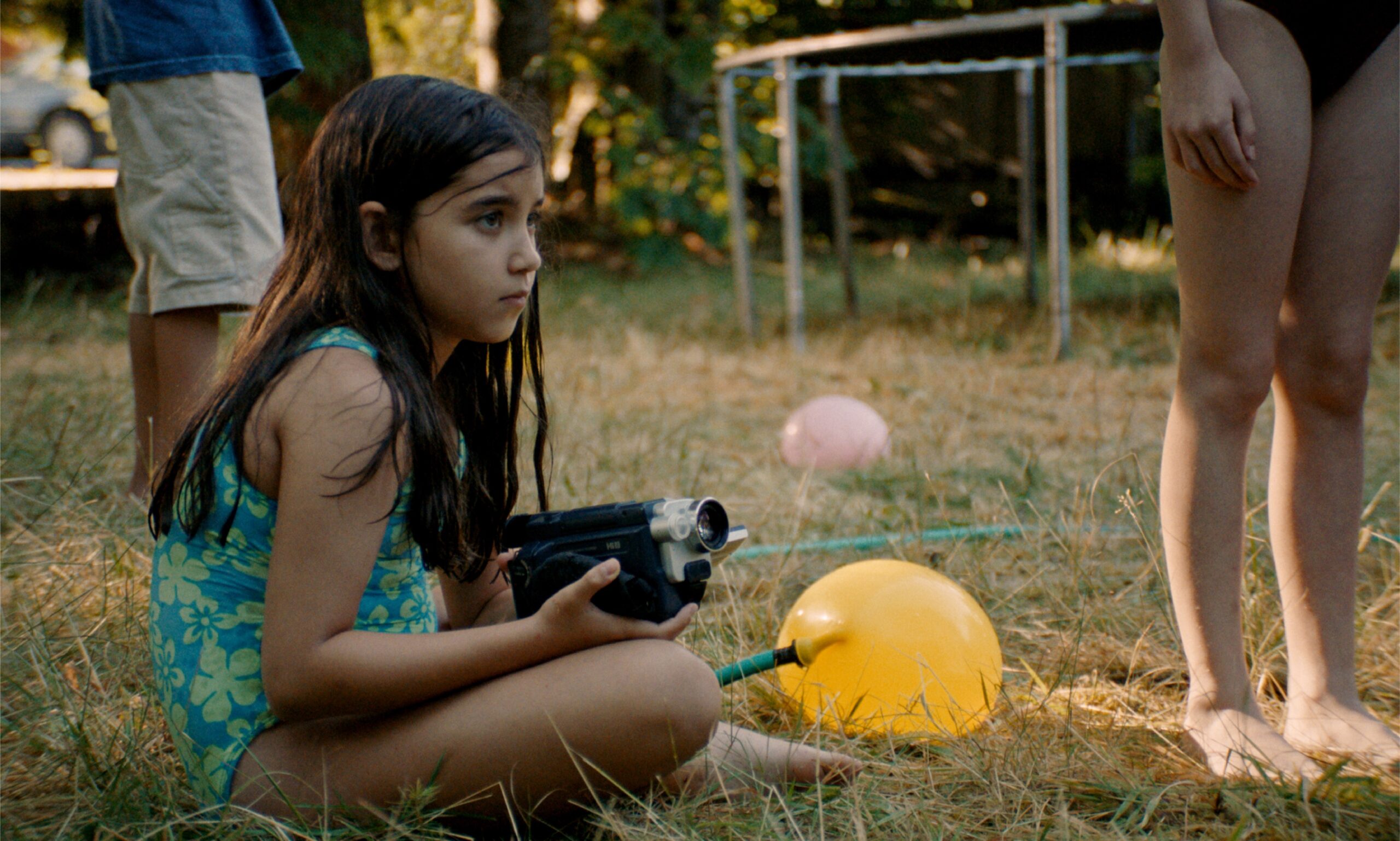 Blue Heron A girl in a patterned swimsuit crouches on dry grass holding a camcorder, looking intently to the side, with balloons, a hose, and other children partially visible around her in a backyard setting.