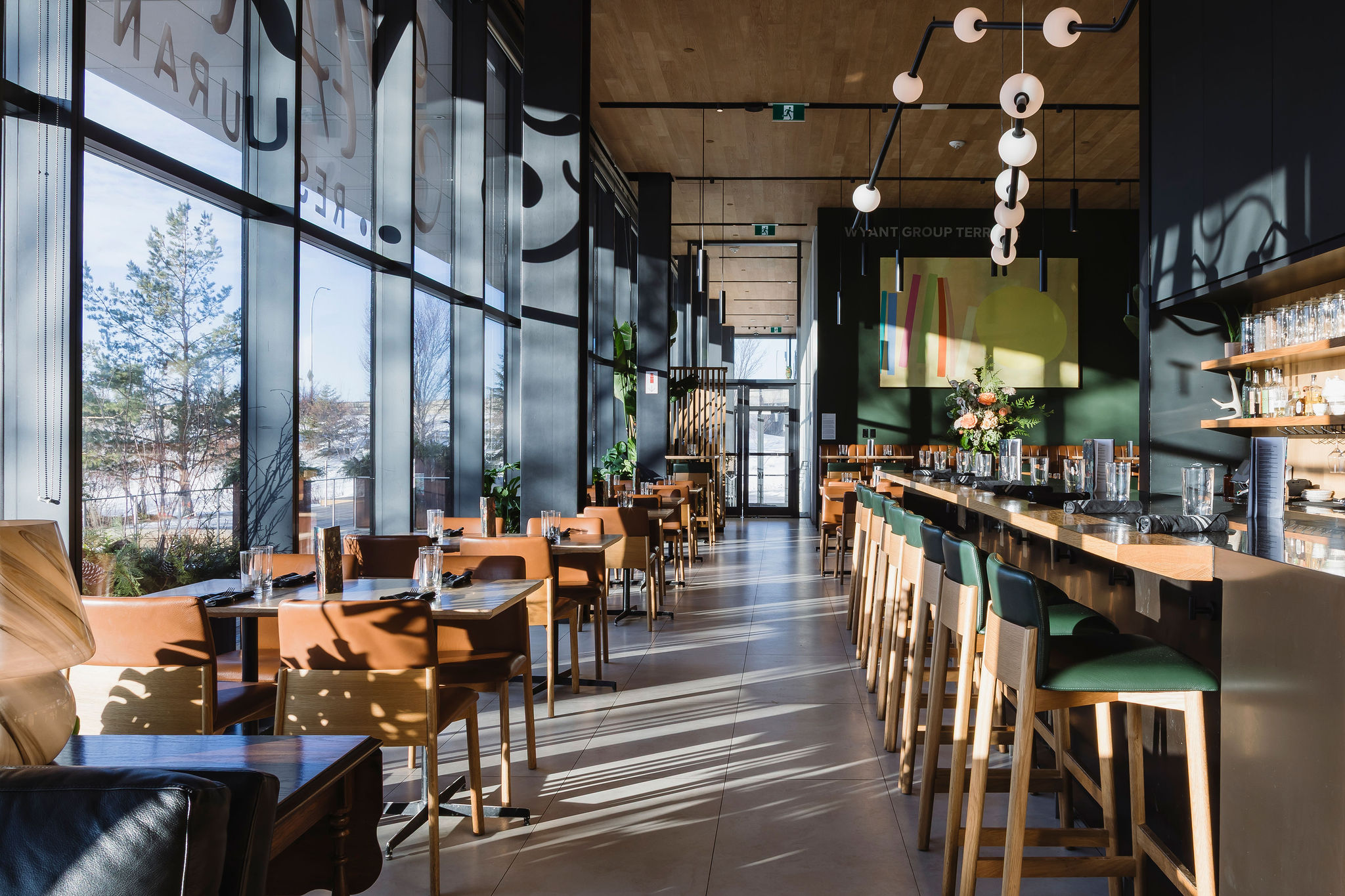 Interior of a modern restaurant with floor-to-ceiling windows, wooden tables and chairs, green upholstered bar seating along a long counter, pendant lighting, and natural light streaming across the floors.