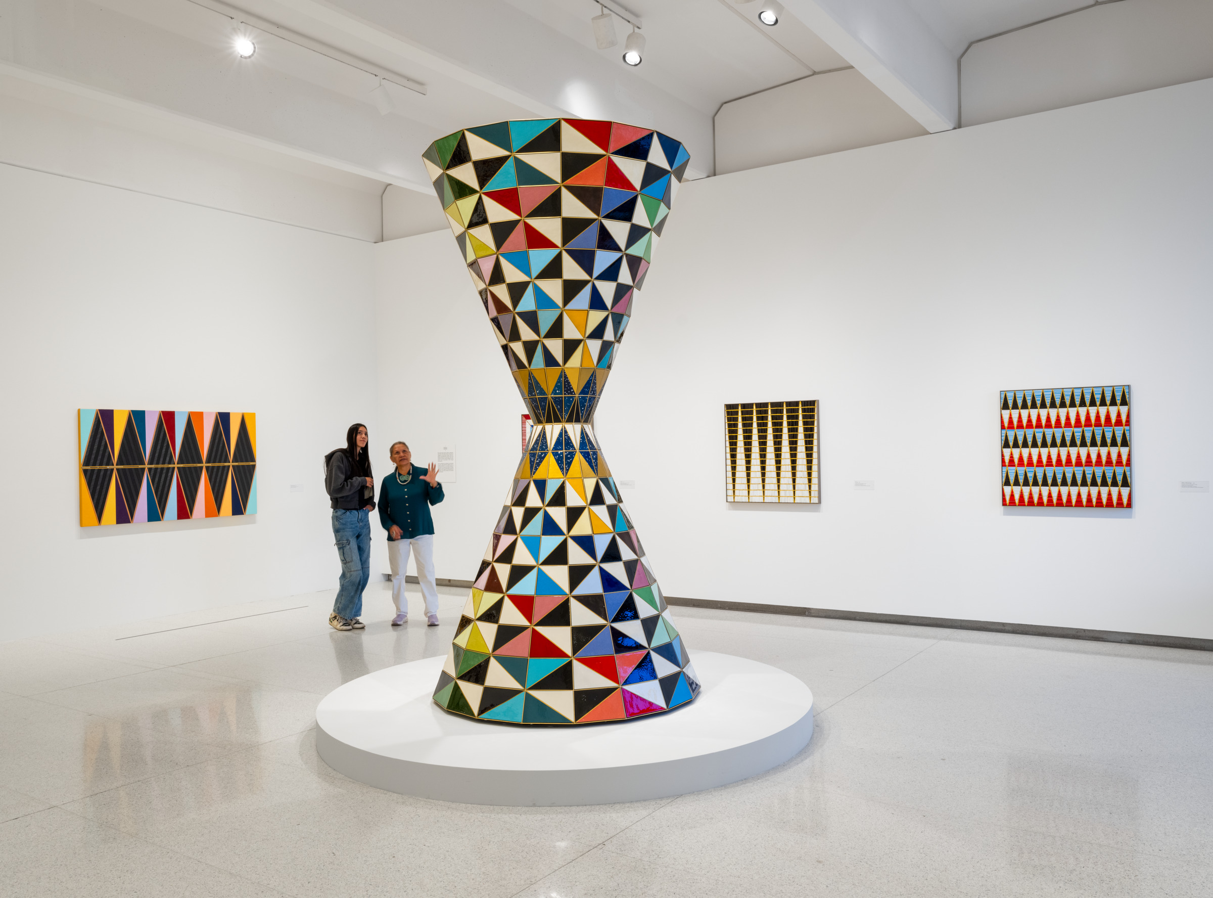 Kameron Herndon for Walker Art Center A large hourglass-shaped sculpture covered in colorful geometric tiles — triangles in red, blue, green, yellow, black, and white — stands on a circular white platform in a bright gallery space. Two visitors stand nearby, one gesturing as if explaining the work. Abstract geometric paintings in similar bold patterns hang on the surrounding white walls.