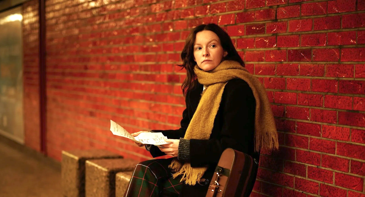 MEASURES A woman sits against a red brick wall, wearing a dark coat and a tan scarf. She holds a few sheets of paper and looks off to the side, with a bag resting beside her on the bench.