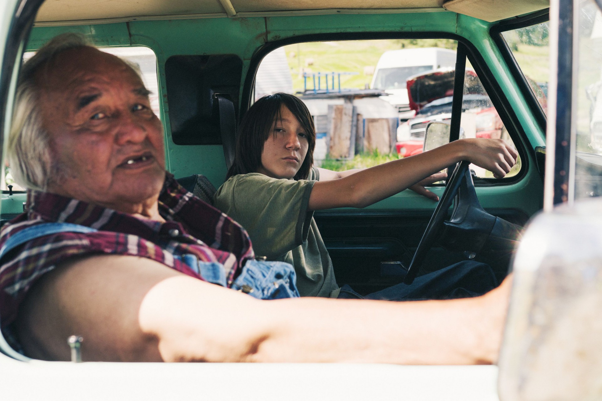 Portraits from a Fire An elderly man in a plaid shirt and overalls sits in the passenger seat of a pickup truck, while a teenage boy leans on the steering wheel in the driver’s seat, both looking toward the camera, with a rural yard and parked vehicles visible outside.