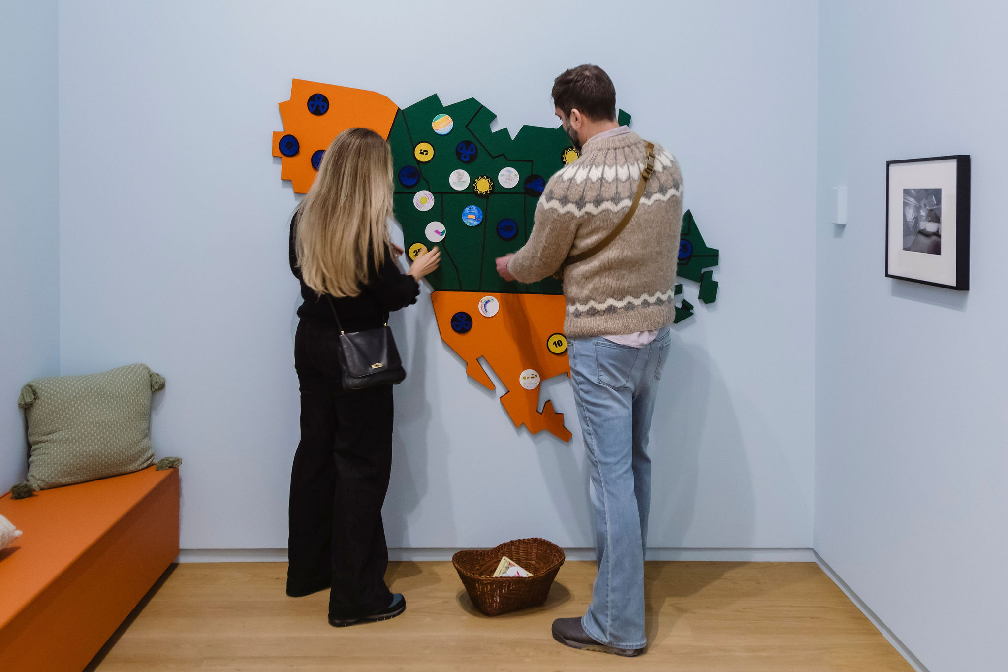 Two visitors interact with a colorful, wall-mounted map-like installation, placing circular pieces onto its surface in a gallery space.