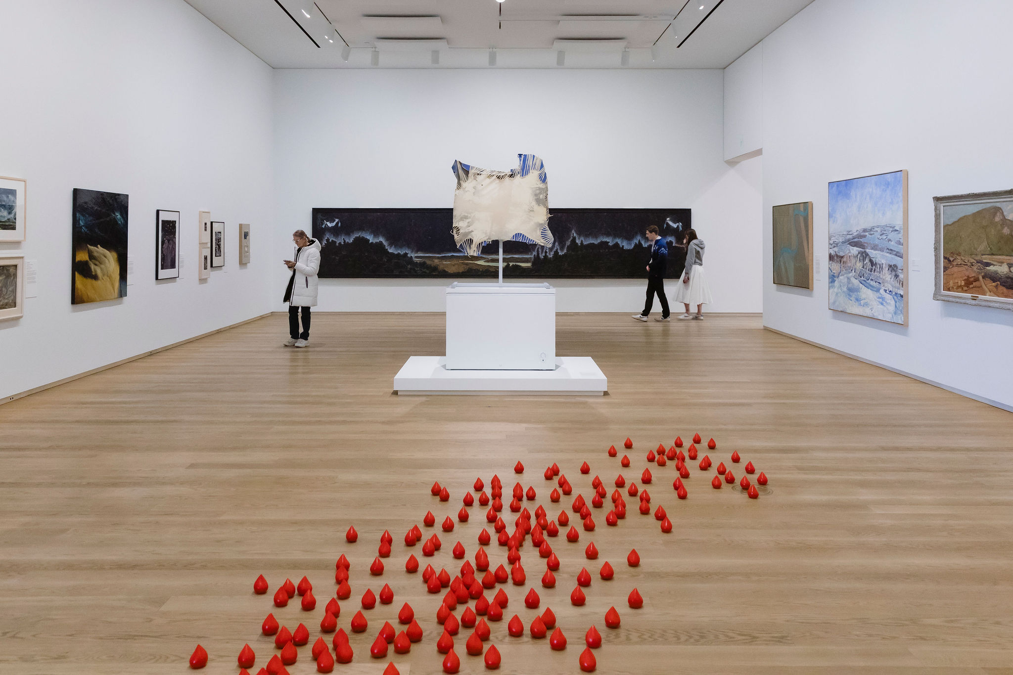 Visitors explore a spacious gallery featuring paintings and sculptures, with a central suspended artwork and a cluster of small red sculptural forms arranged across the floor in the foreground.