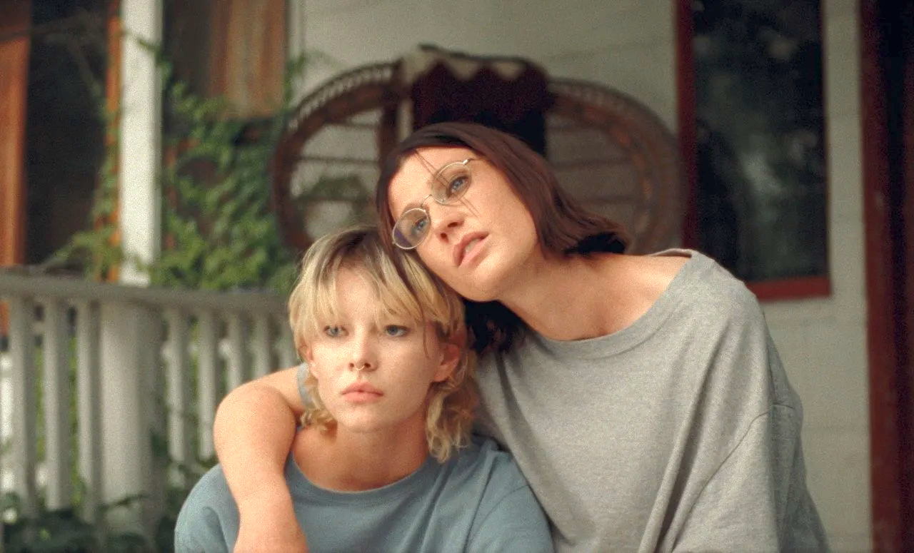 UBB A person with glasses rests her head gently against a younger blonde person, arm wrapped around the other's shoulders. Both wear casual t-shirts and look forward with calm, slightly pensive expressions. They sit together on a porch with greenery and wooden details softly blurred in the background.