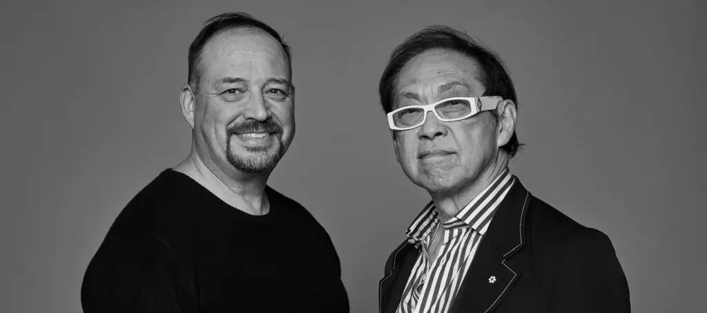 architects banner portrait new building 1024×454.jpg (1) 1 Black and white portrait of two smiling men standing side by side. One wears a dark sweater, the other wears a striped shirt with dark jacket and white-framed glasses.
