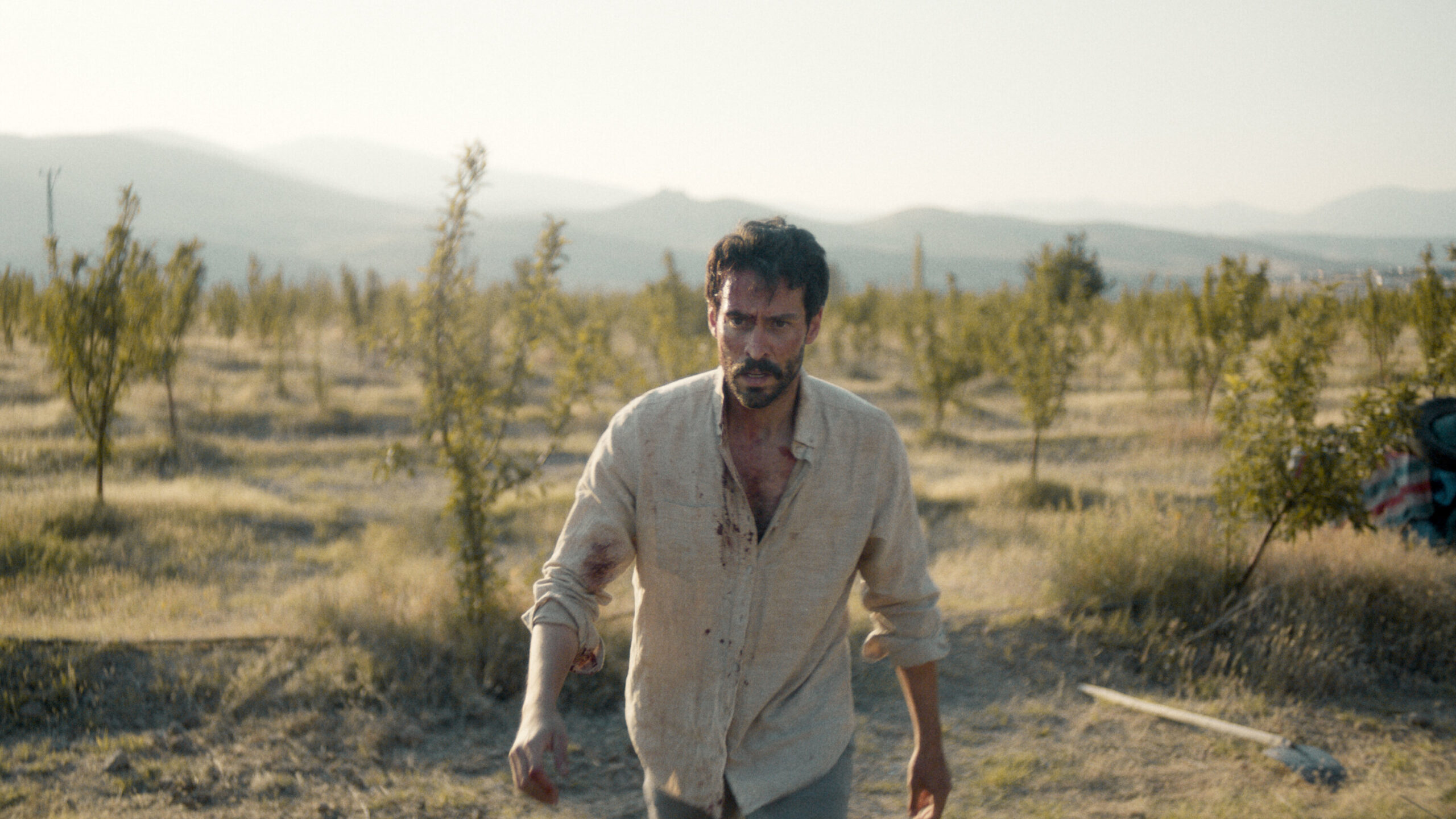 thingsyoukill A disheveled man with bloodstains on his shirt walks through a dry field of small trees, with mountains in the distance under a hazy sky.