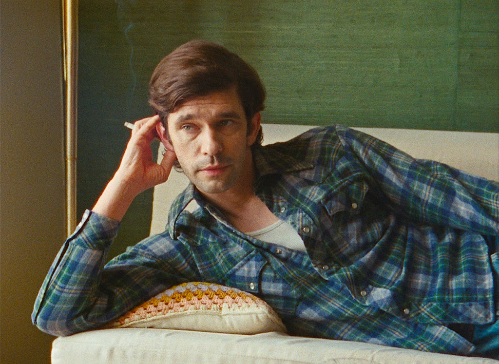 Man with dark hair reclining on a couch, wearing a blue plaid shirt and white undershirt, looking off to the side against a green wall background.