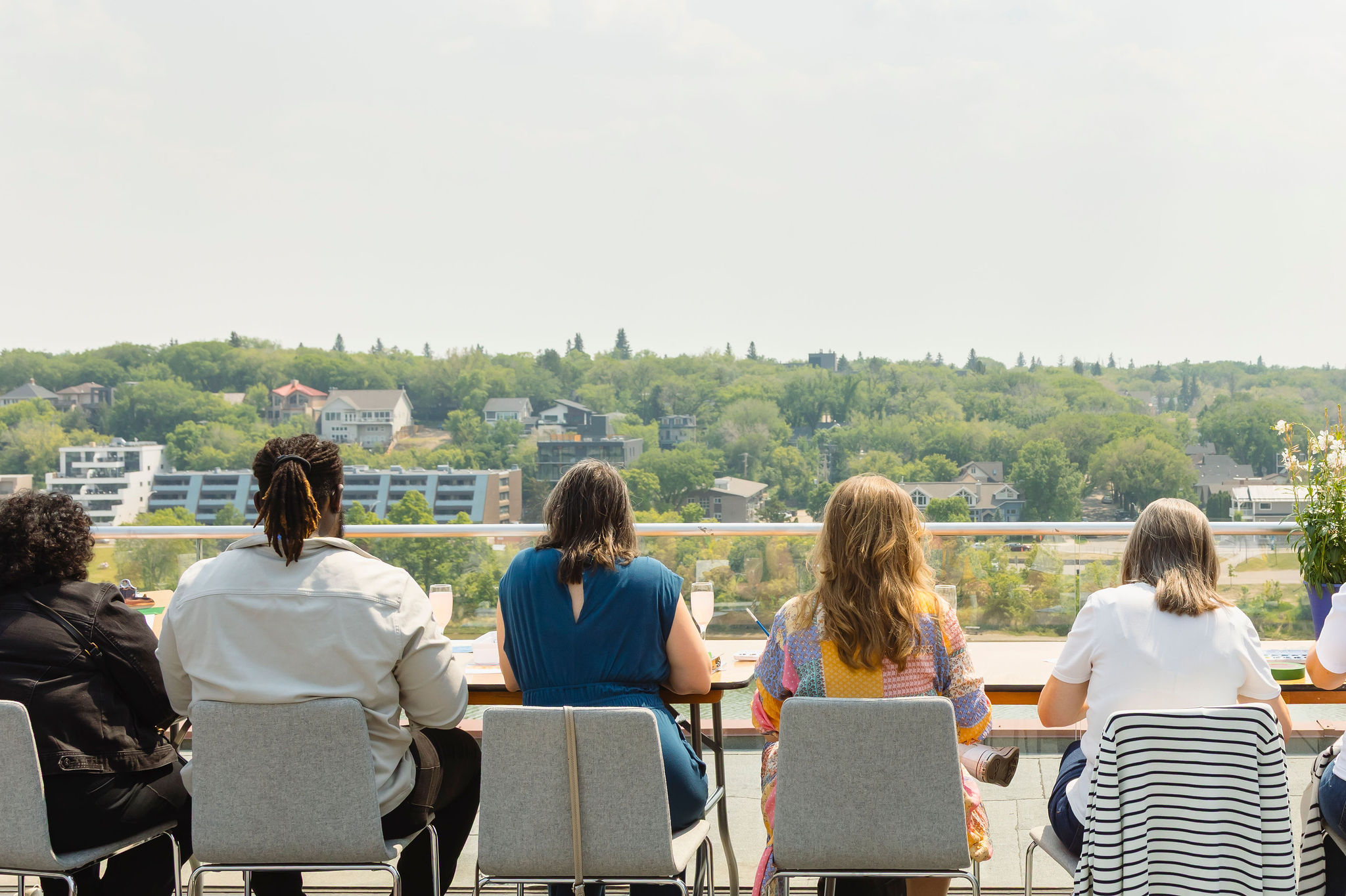 Six participants sit with their backs to the camera at a long table on Remai Modern's rooftop patio, painting watercolours while looking out over a panoramic view of Saskatoon's South Saskatchewan River valley and tree-covered riverbank neighbourhoods on a sunny day.