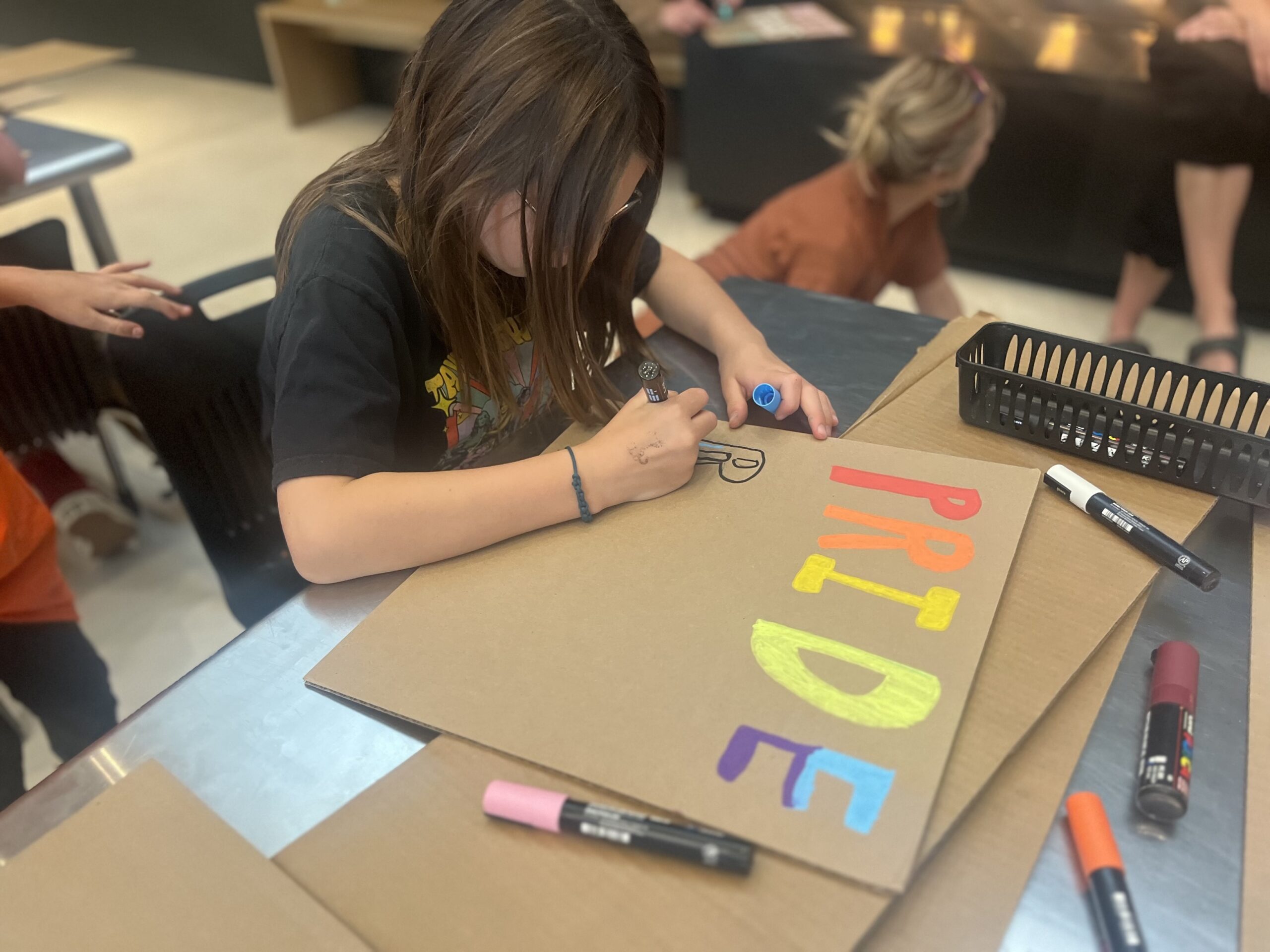 A child sits at a table focused on decorating a large piece of cardboard with colorful letters spelling “PRIDE.” They are using markers and paint pens while other children and adults work on projects in the background, creating a creative classroom or workshop atmosphere.