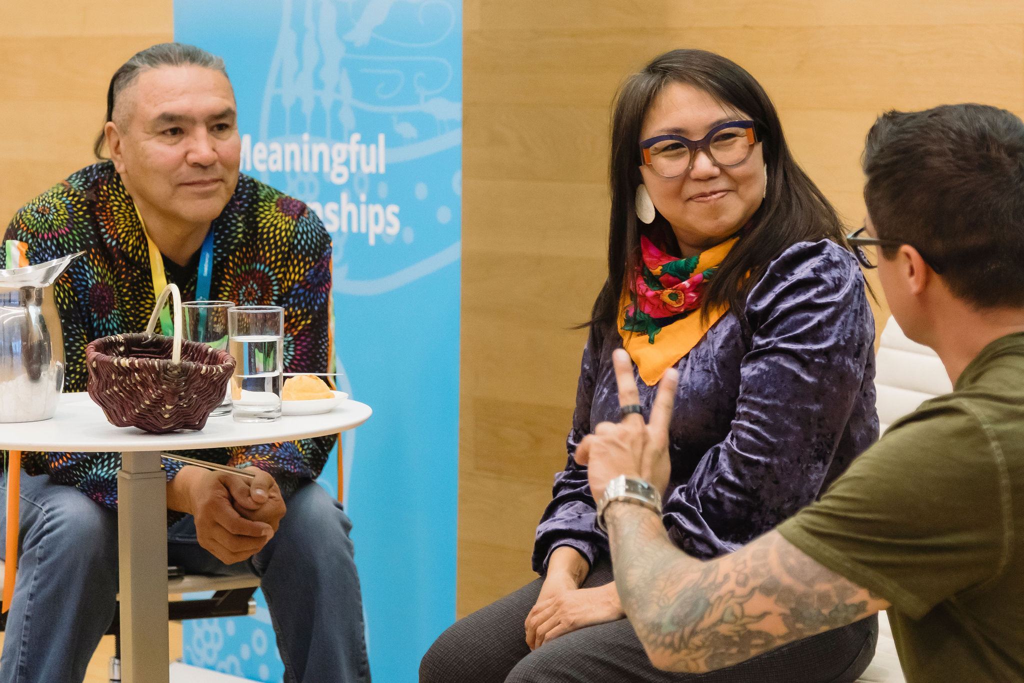 Remai FiresideLucyTulugarjuk 039 Three people participate in a seated discussion at an indoor event. On the left, a person in a brightly patterned shirt sits beside a small round table holding a water pitcher, glasses, and a woven basket. On the right, a person wearing glasses, a purple velvet top, and a colorful scarf smiles while listening to another participant in the foreground who is gesturing with their hand. A blue banner and wood-panelled wall form the background.