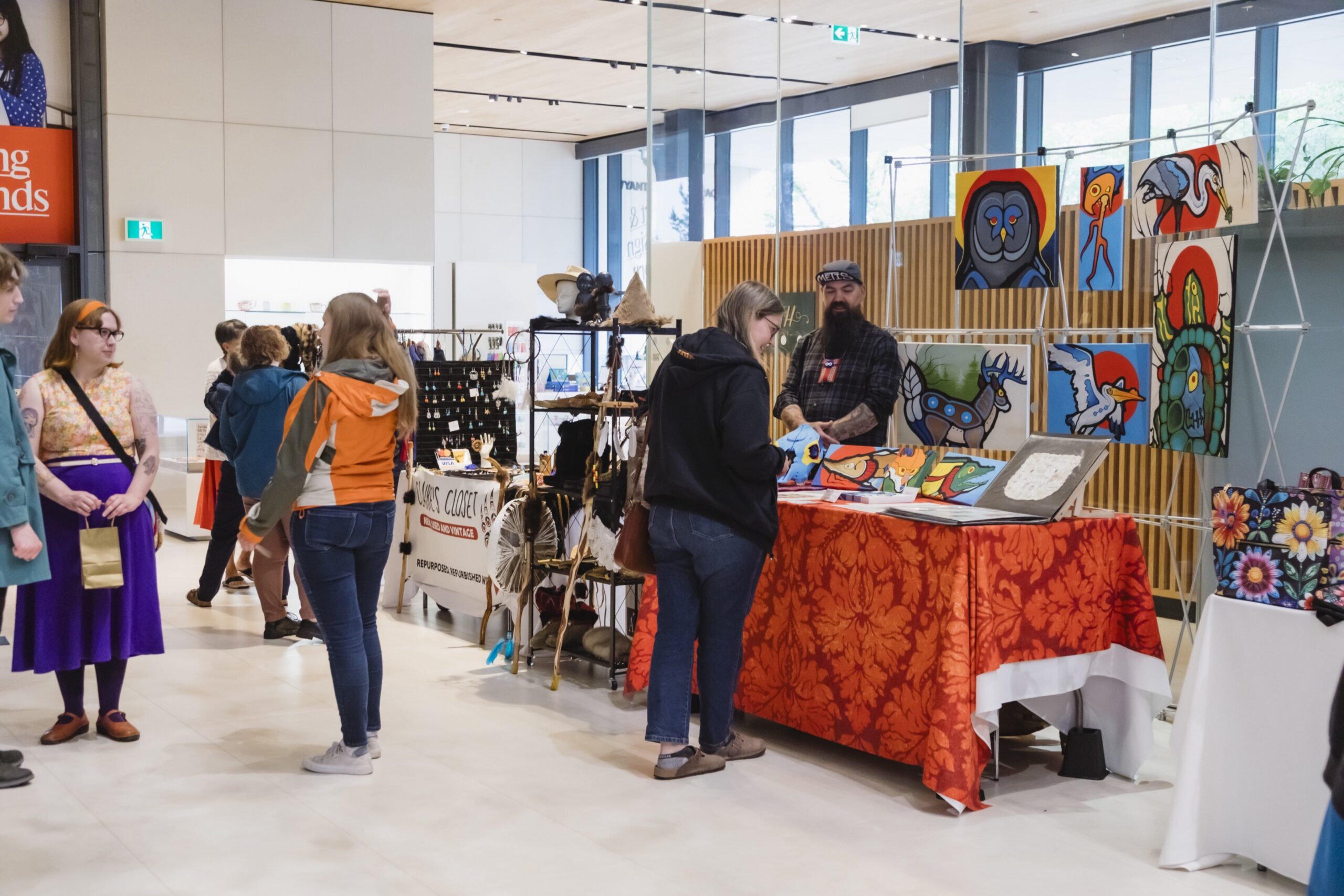 Remai MétisMarket2025 0009 Visitors browse artist and artisan booths at an indoor Métis market, featuring colorful Indigenous artwork, handmade goods, and crafts displayed throughout a bright gallery space.