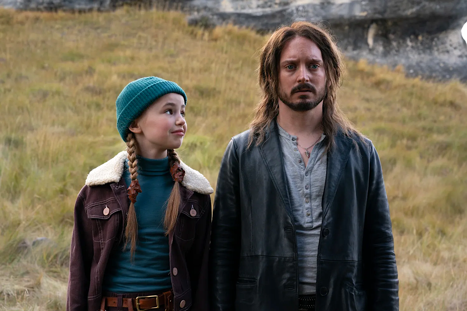 A young girl in a teal knit hat smiles up at a long-haired man in a black leather jacket as they stand side by side in a grassy, rocky landscape.