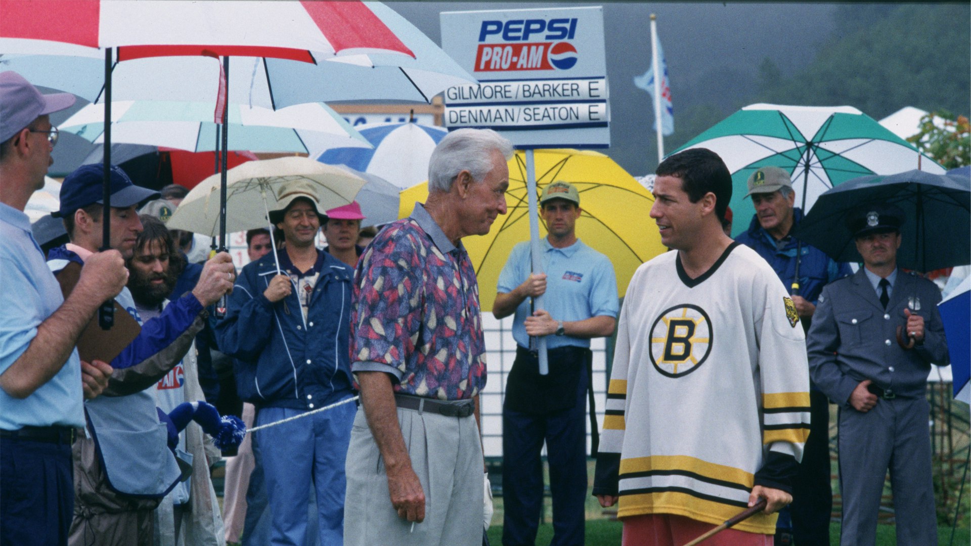 A man in a hockey jersey talks with an older man at a rainy golf tournament, surrounded by spectators holding colorful umbrellas near a leaderboard.