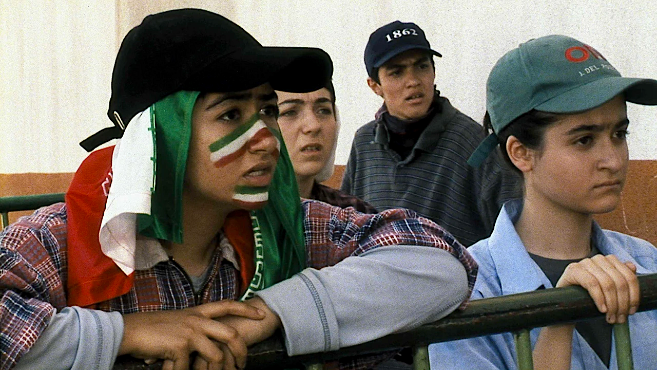 A group of young spectators lean on a railing, watching intently; one wears face paint and a flag draped over their head in support of a team.