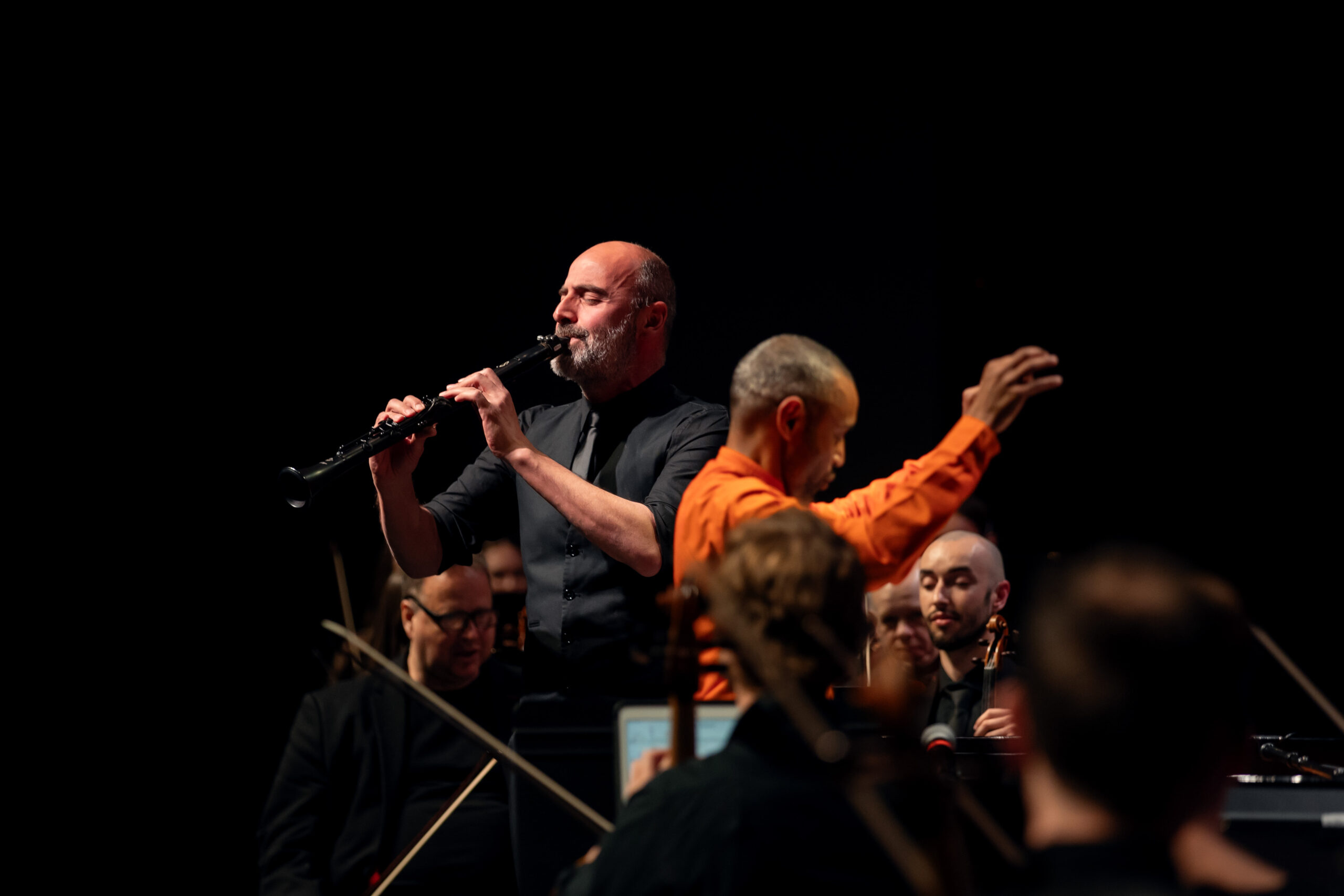 A clarinet soloist performs alongside an orchestra, while a conductor in an orange shirt leads the ensemble on stage.